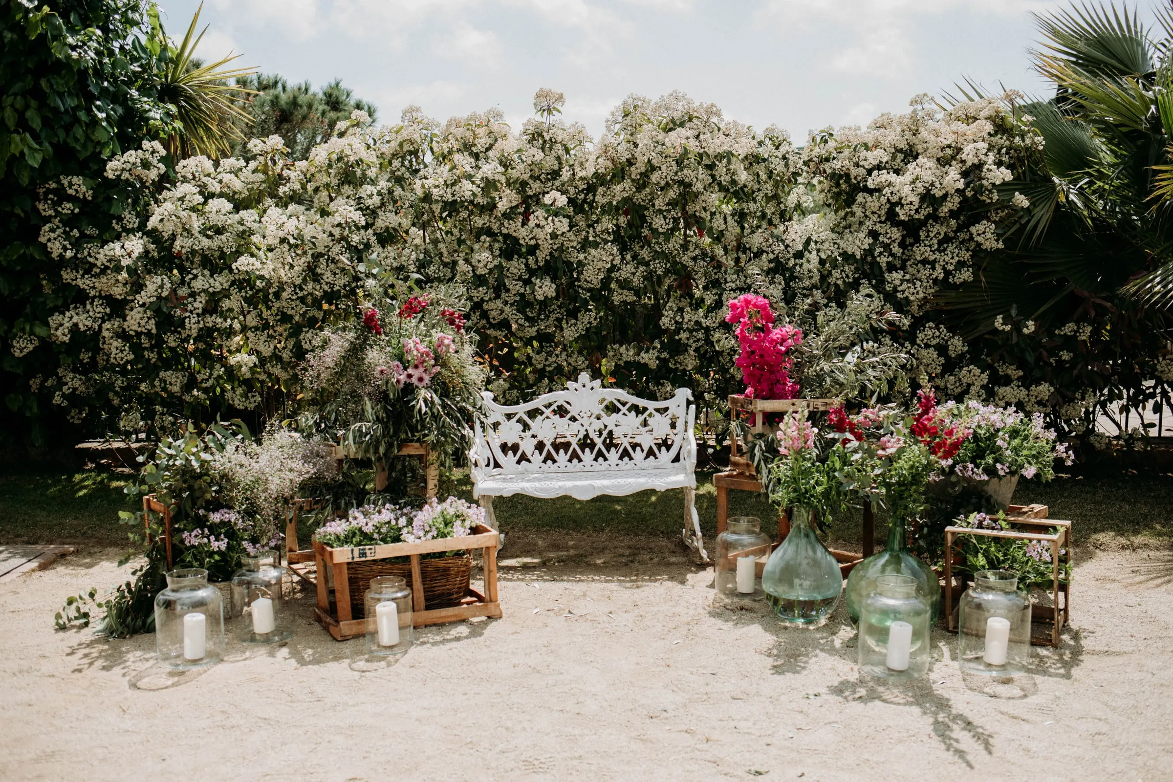 Un banco de forja blanco entre flores rosas y rojas y jarrones de cristal ante un seto en flor, en una masía de Girona