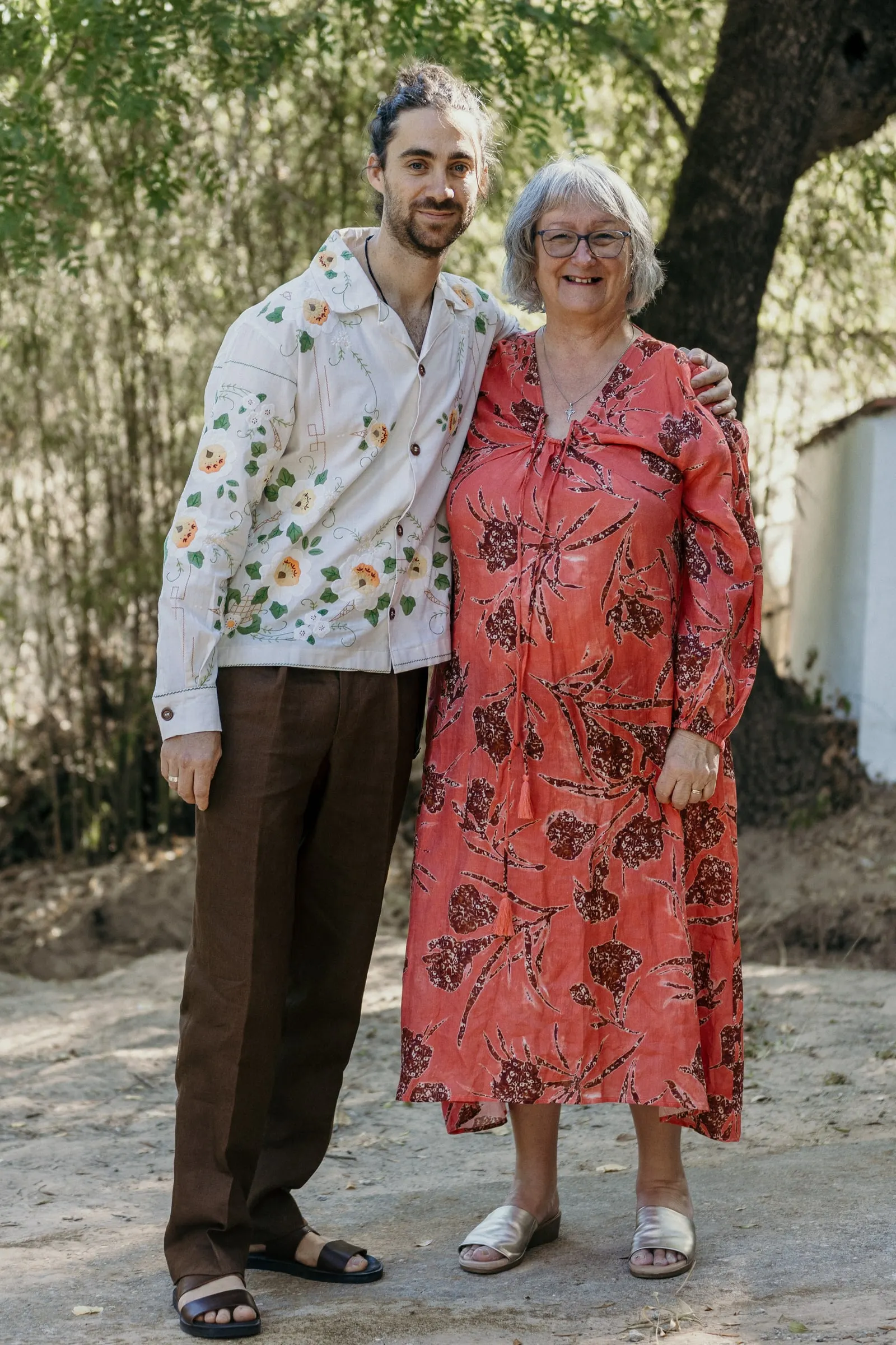 El novio con camisa bordada de flores posa con su madre a la sombra