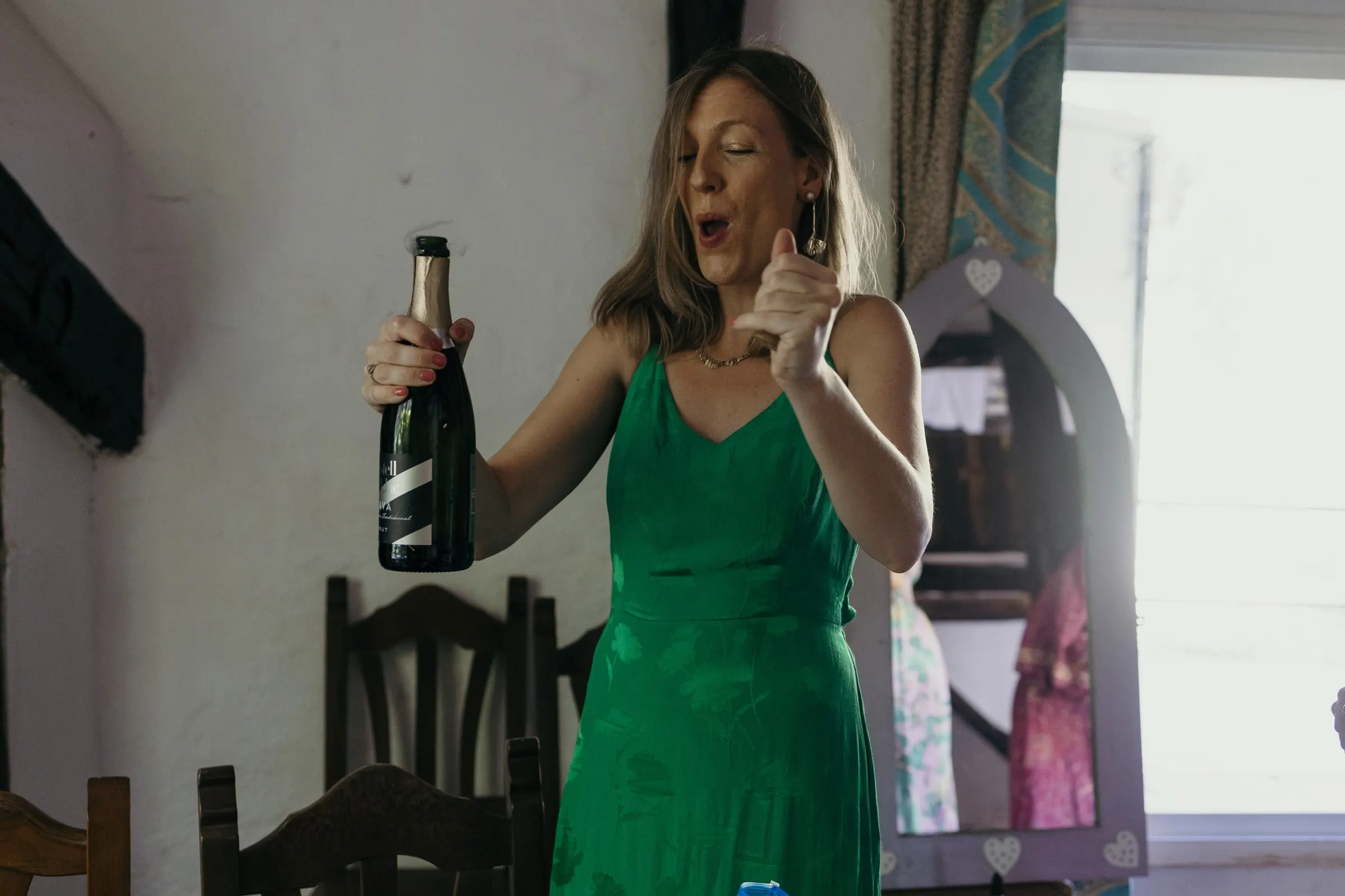 Una amiga de vestido verde abre una botella de cava en la habitación de la novia