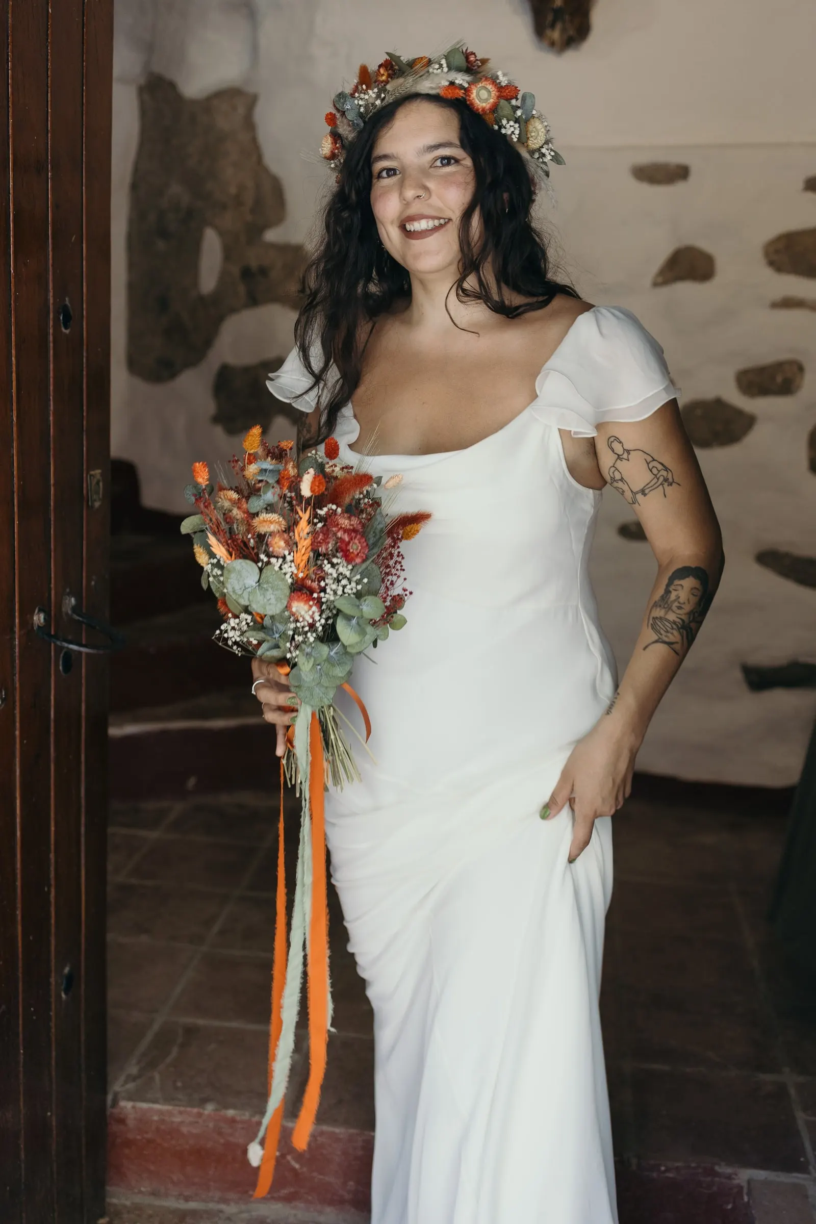 La novia en una puerta con muro de piedra, con un ramo de cintas naranjas