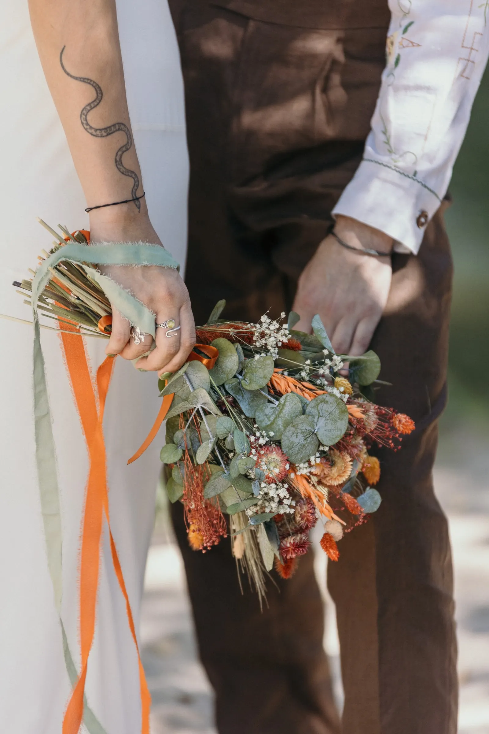 La mano tatuada de la novia con un ramo de flores secas y cintas largas