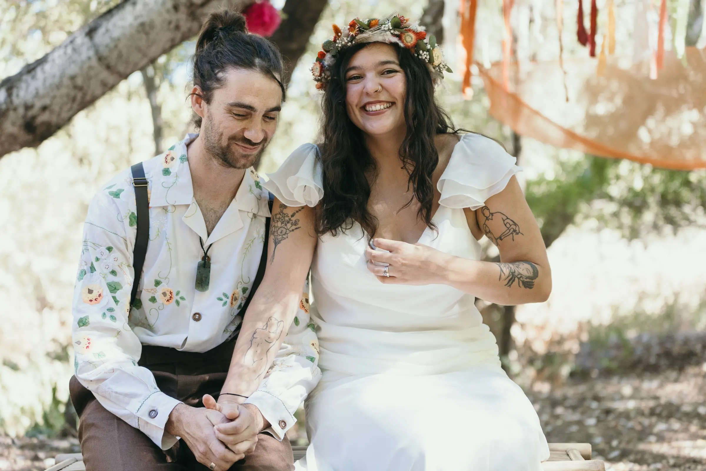 Los novios se ríen sentados en un banco bajo un árbol viejo durante la ceremonia