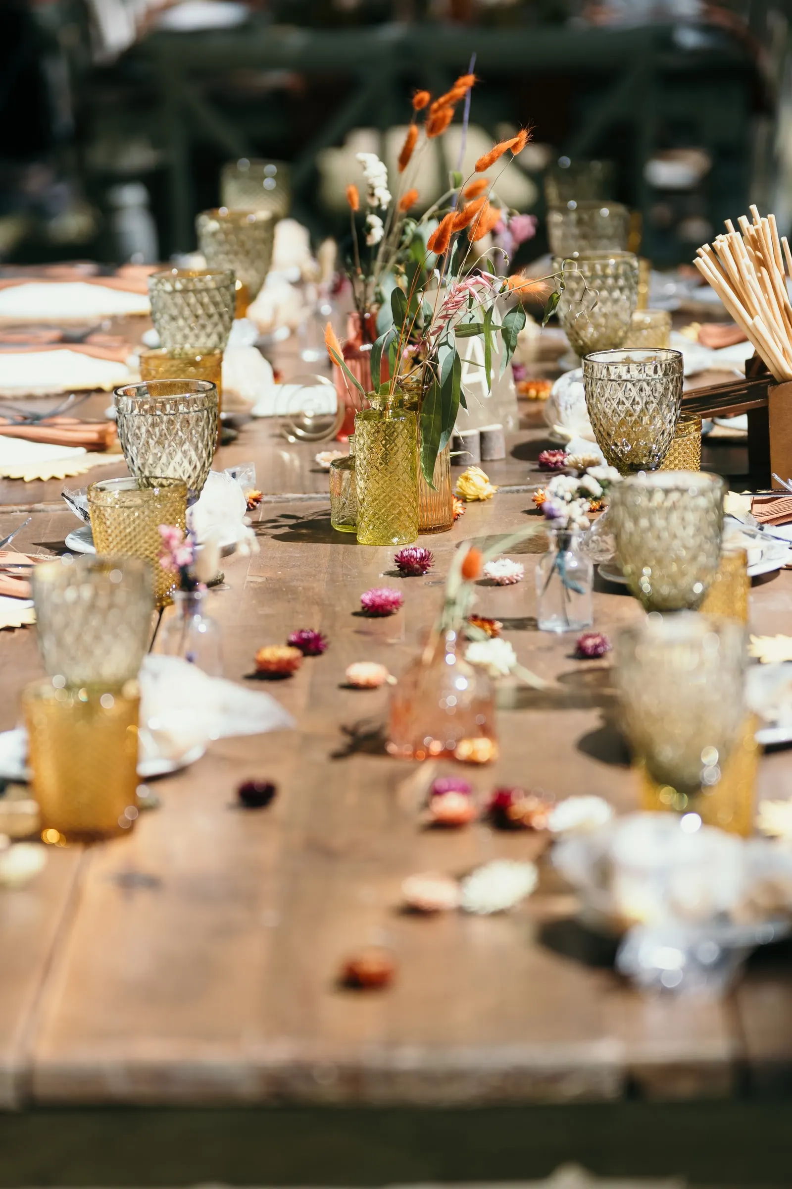 Long wooden dinner table with amber glasses, orange flowers and petals