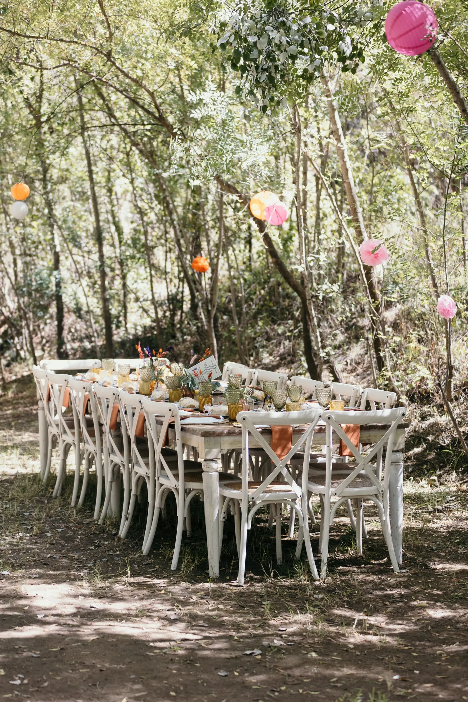 Dinner table with white cross-back chairs under pink paper lanterns in the woods