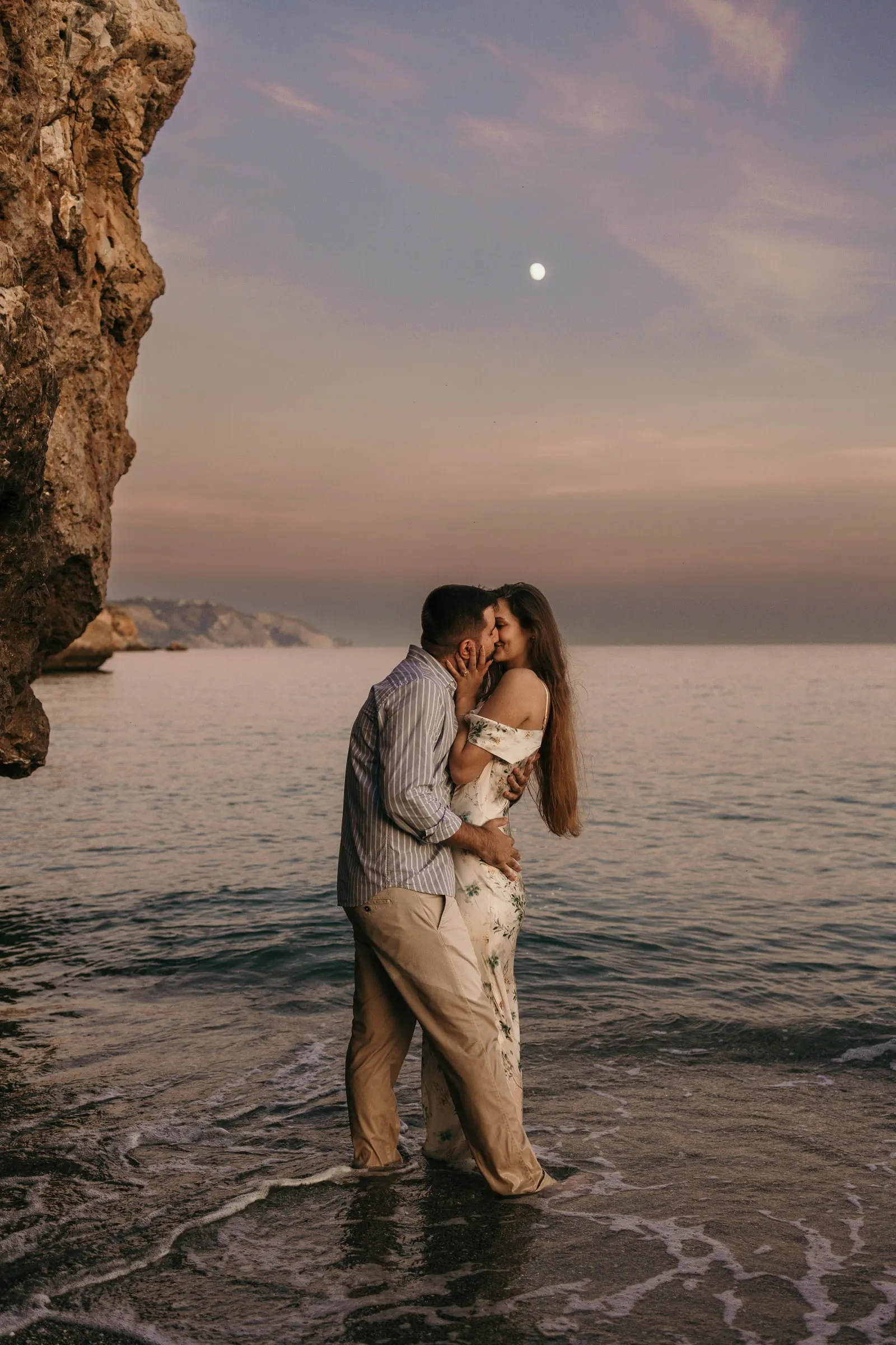 La pareja se besa con el agua por las rodillas al anochecer, con la luna sobre el acantilado, playa de Nerja