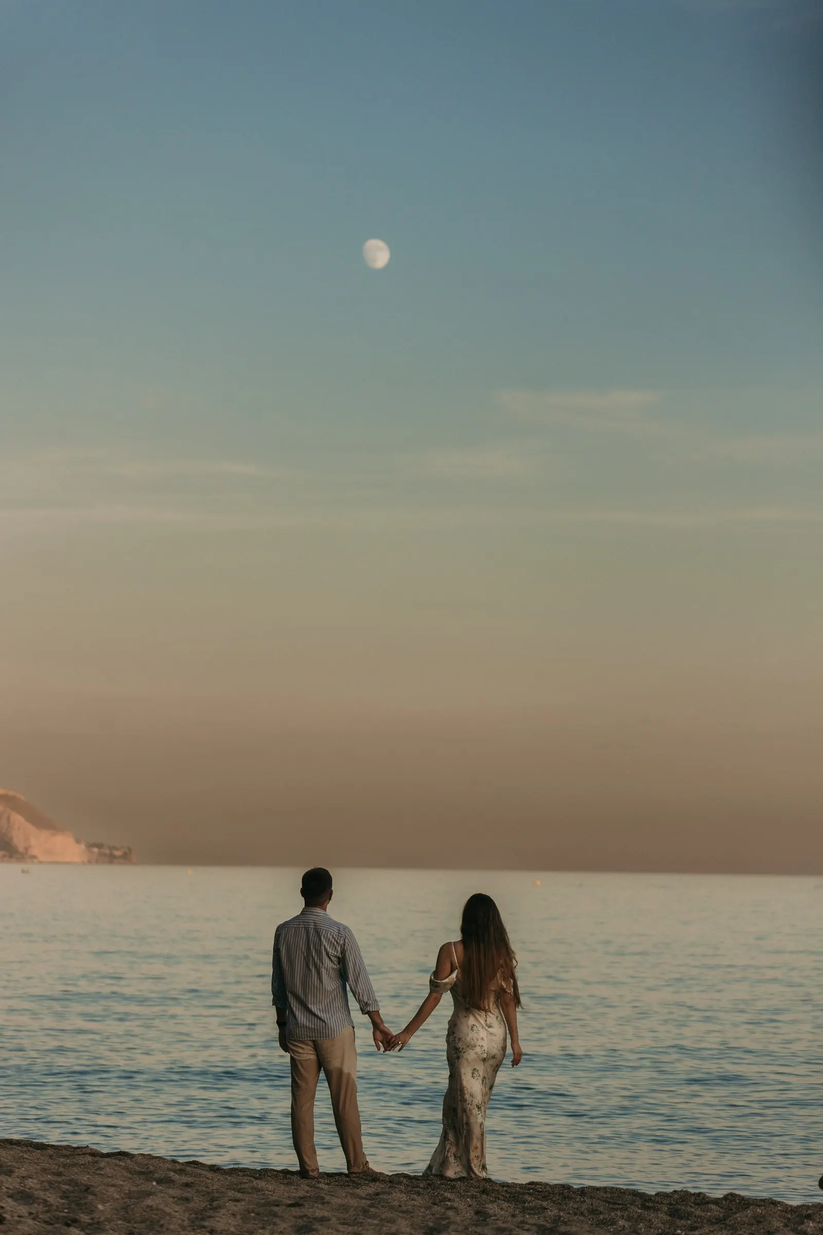 Couple holding hands facing the sea at dusk under a rising moon, on the beach in Nerja