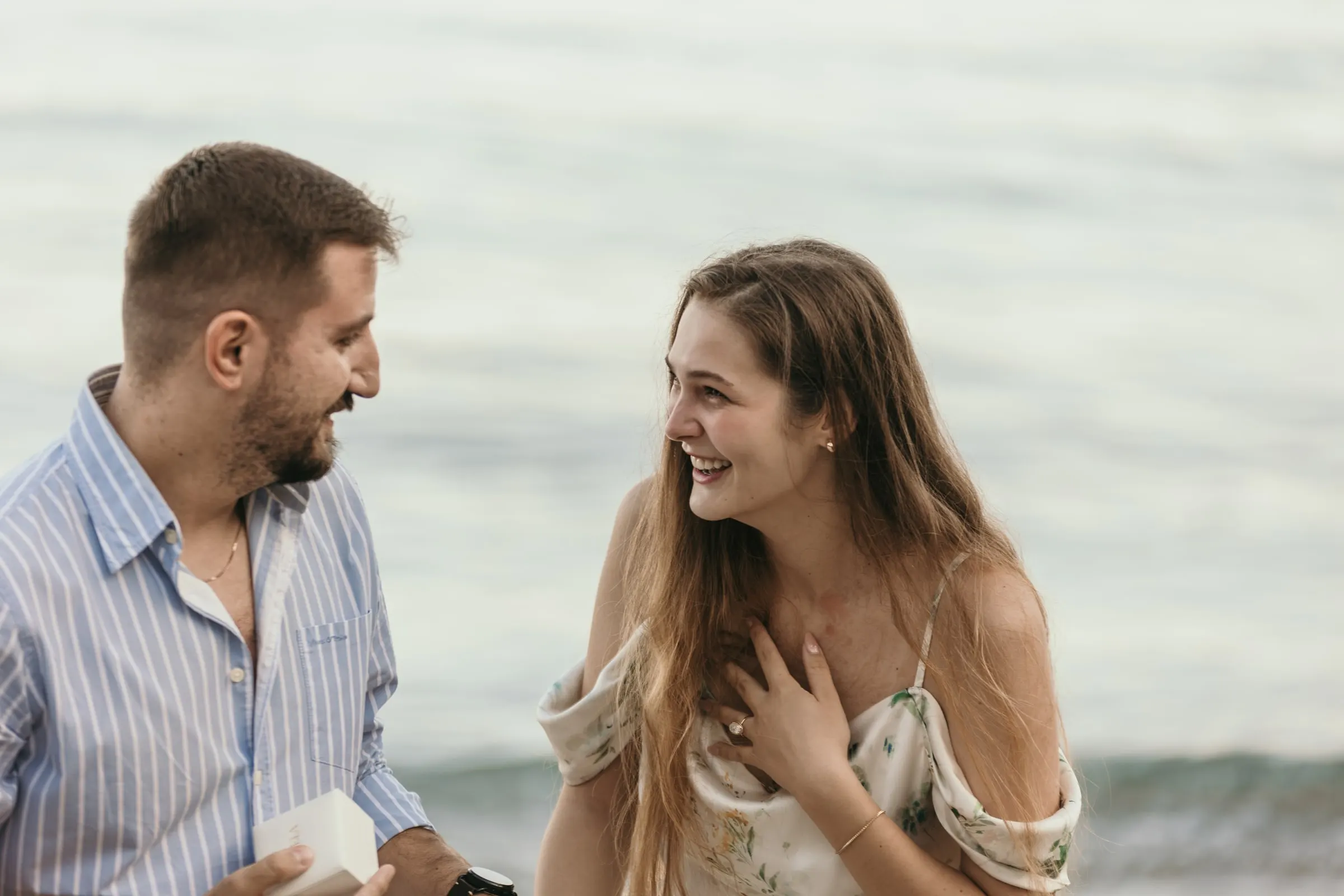 Ella ríe con la mano en el pecho mientras él abre la caja del anillo, en la playa de Nerja