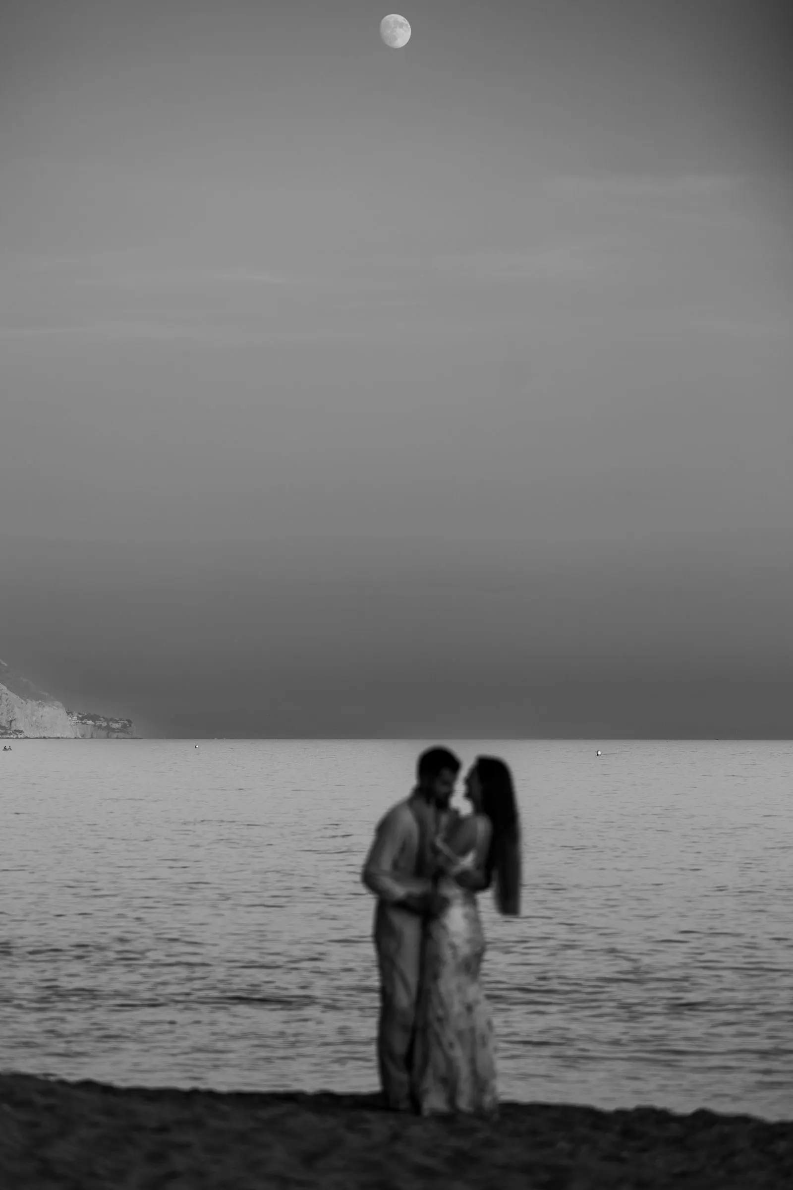 Black and white of the couple small against the sea, the moon above, on the beach in Nerja