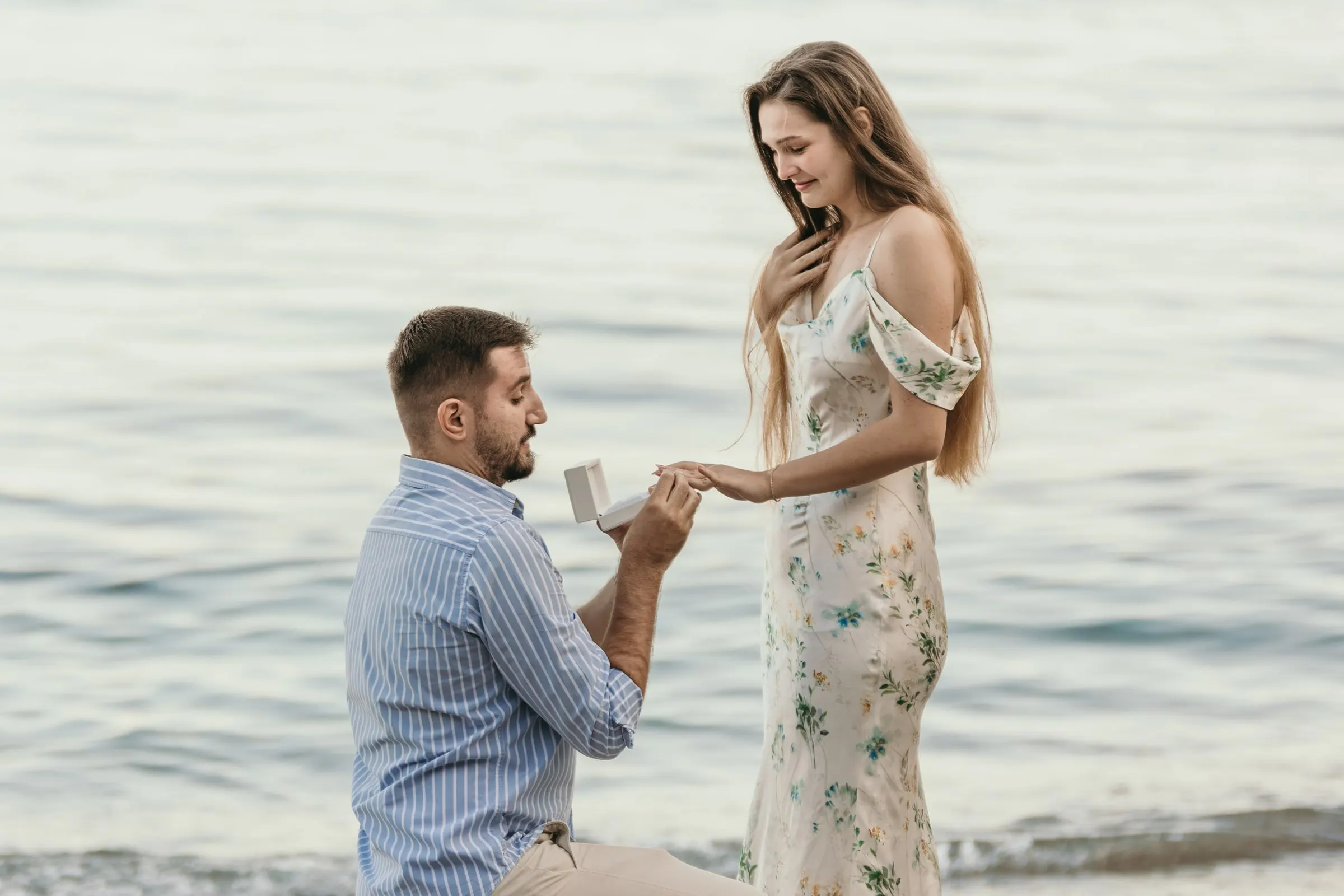 Él le pone el anillo, arrodillado en la orilla, mientras ella sonríe, en la playa de Nerja