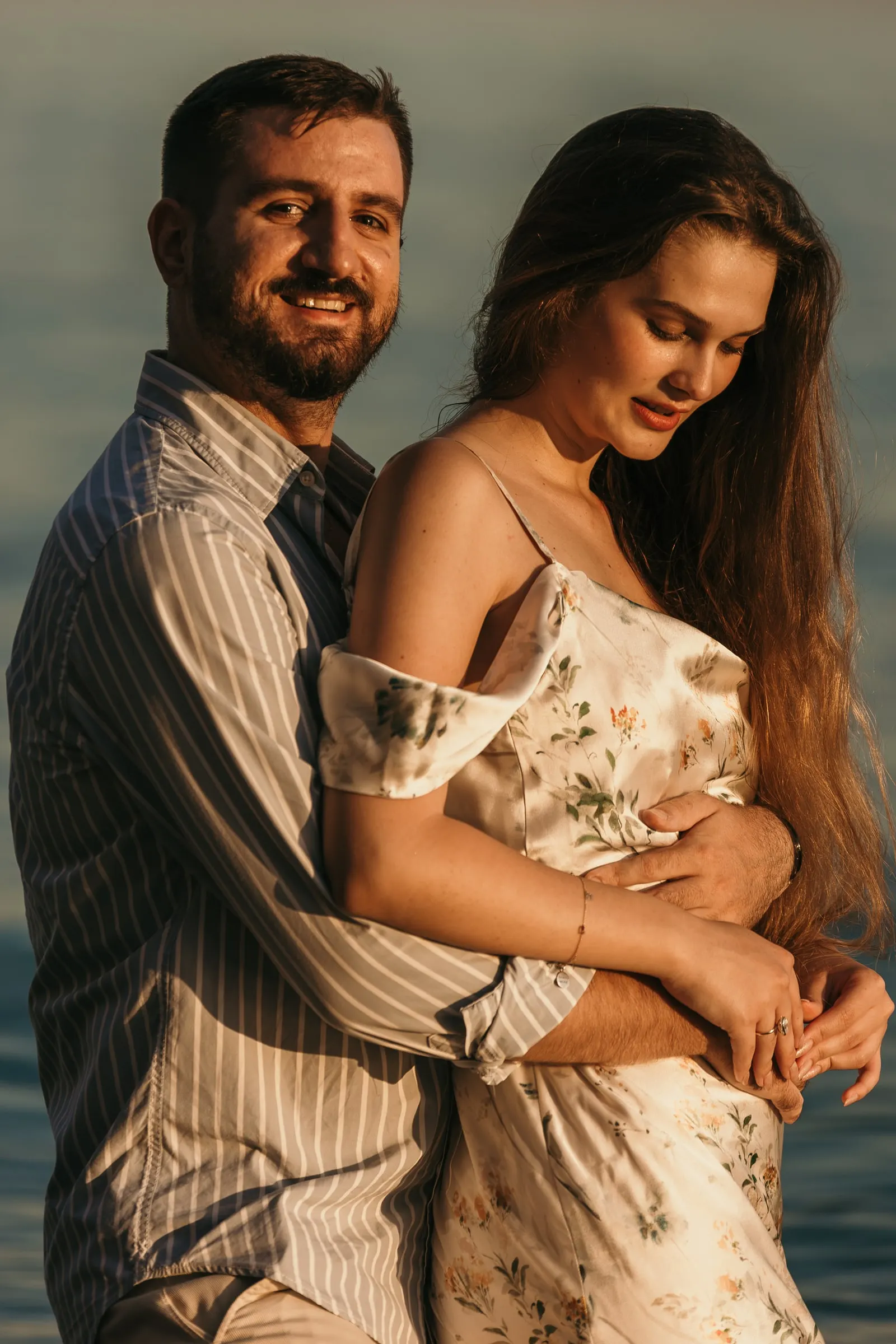 Él la abraza por detrás y los dos sonríen con la luz cálida de la tarde, playa de Nerja