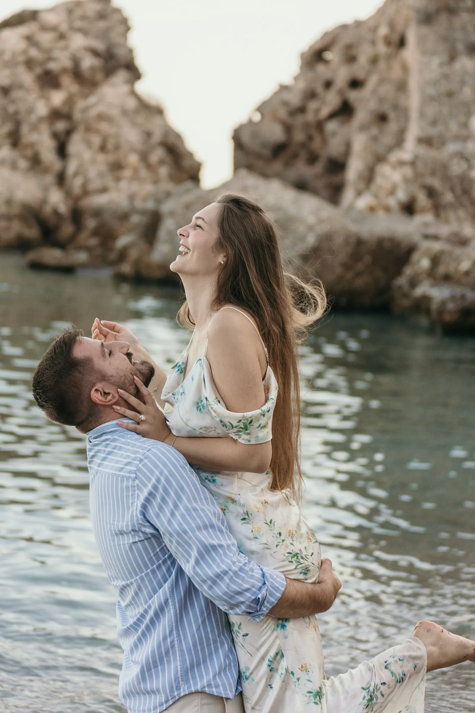 Ella echa la cabeza atrás riendo mientras él la levanta junto a las rocas, playa de Nerja