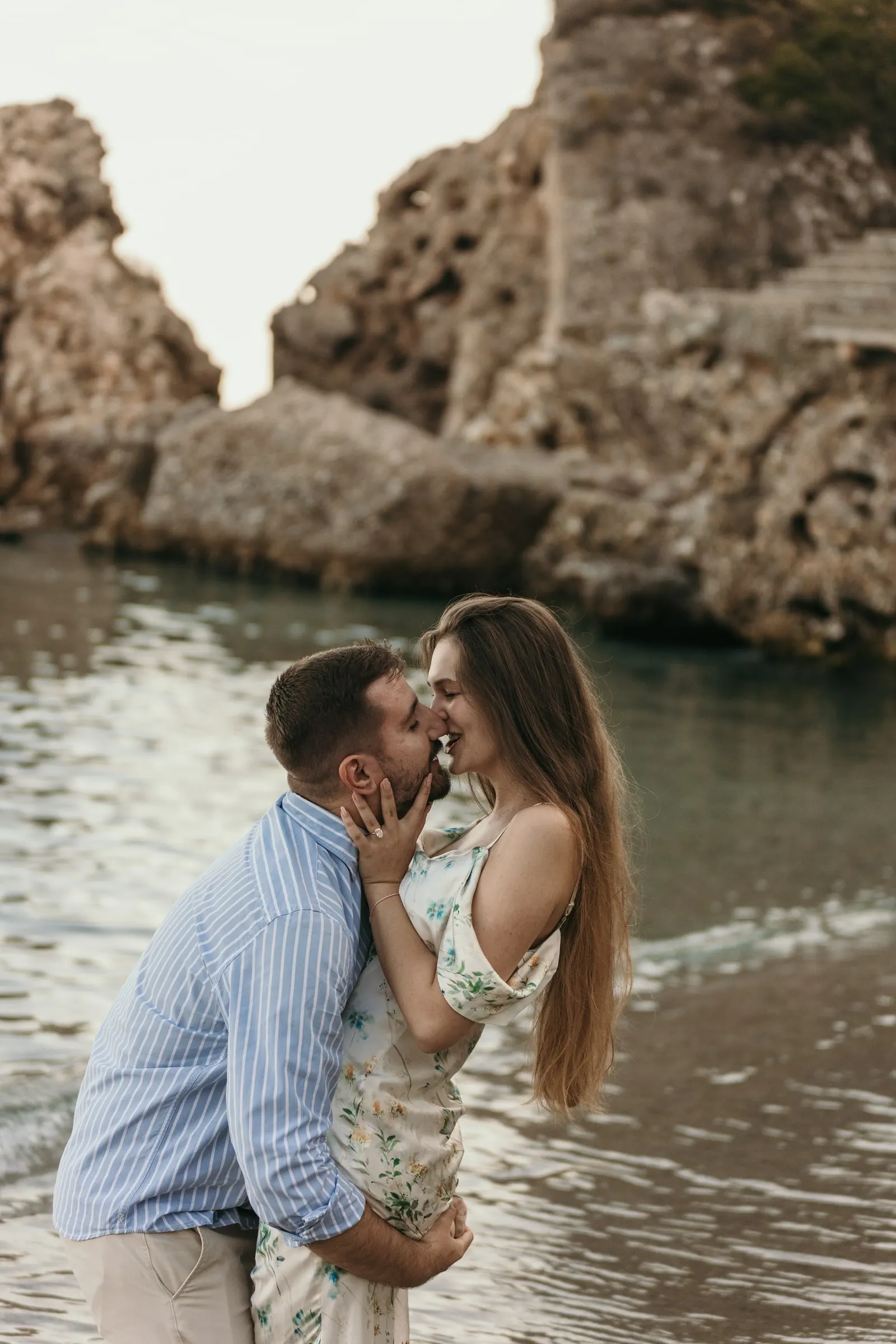 Él la levanta para besarla en el agua, frente a la cala rocosa, en la playa de Nerja