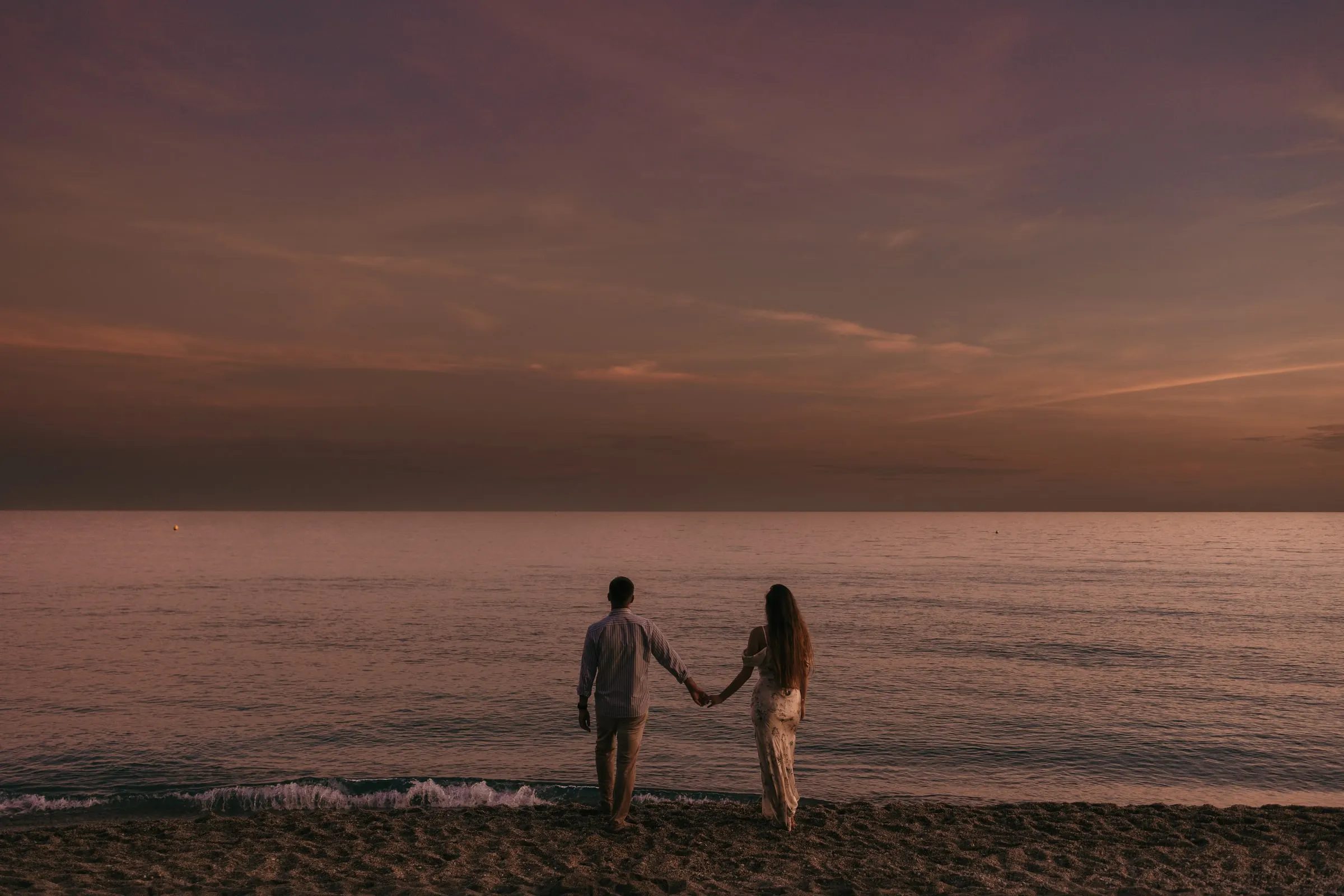 Couple holding hands facing the sea under a violet sunset sky, on the beach in Nerja