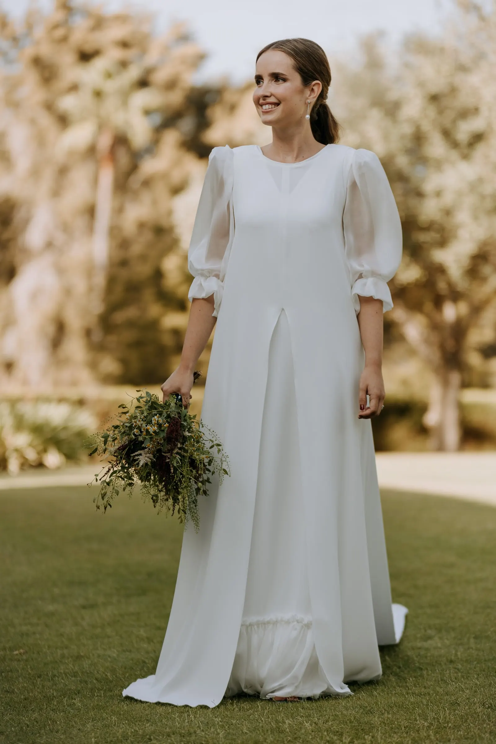 Bride smiling at the camera with her wild-flower bouquet among the trees of the garden