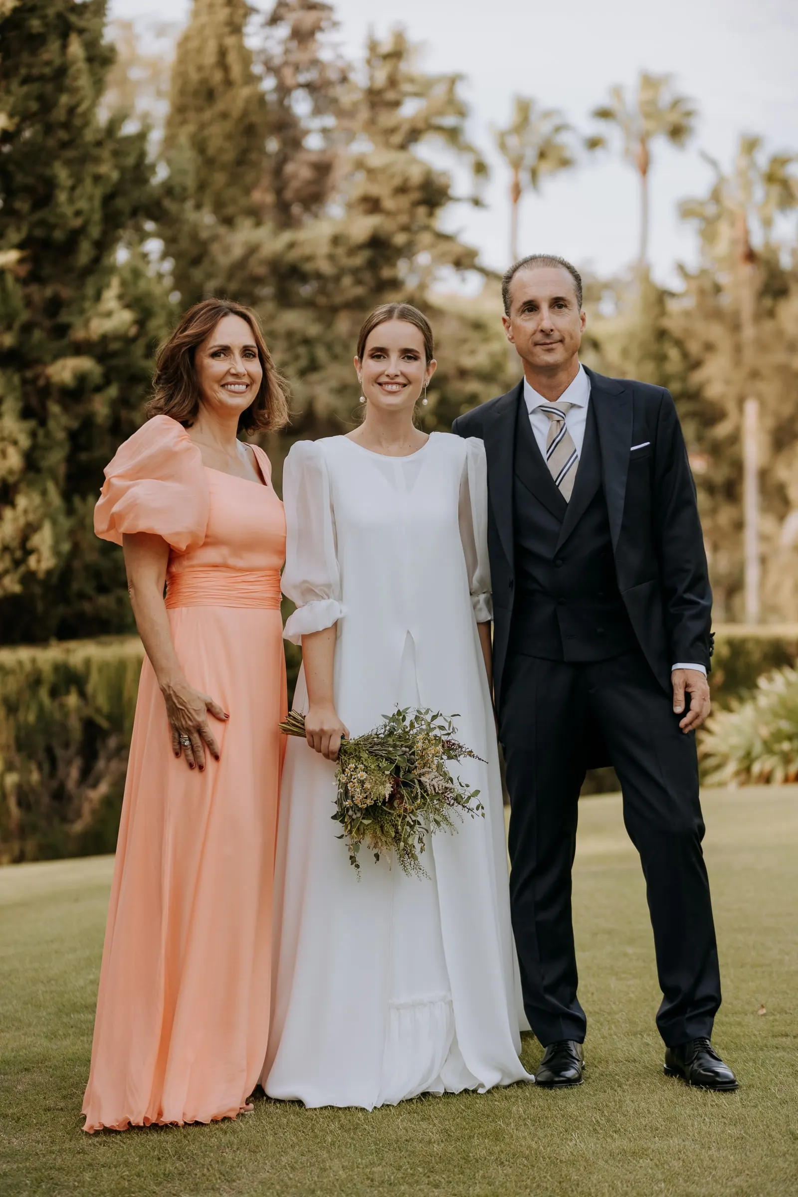 Bride with a woman in a peach gown and a man in a morning suit on the lawn of the family home