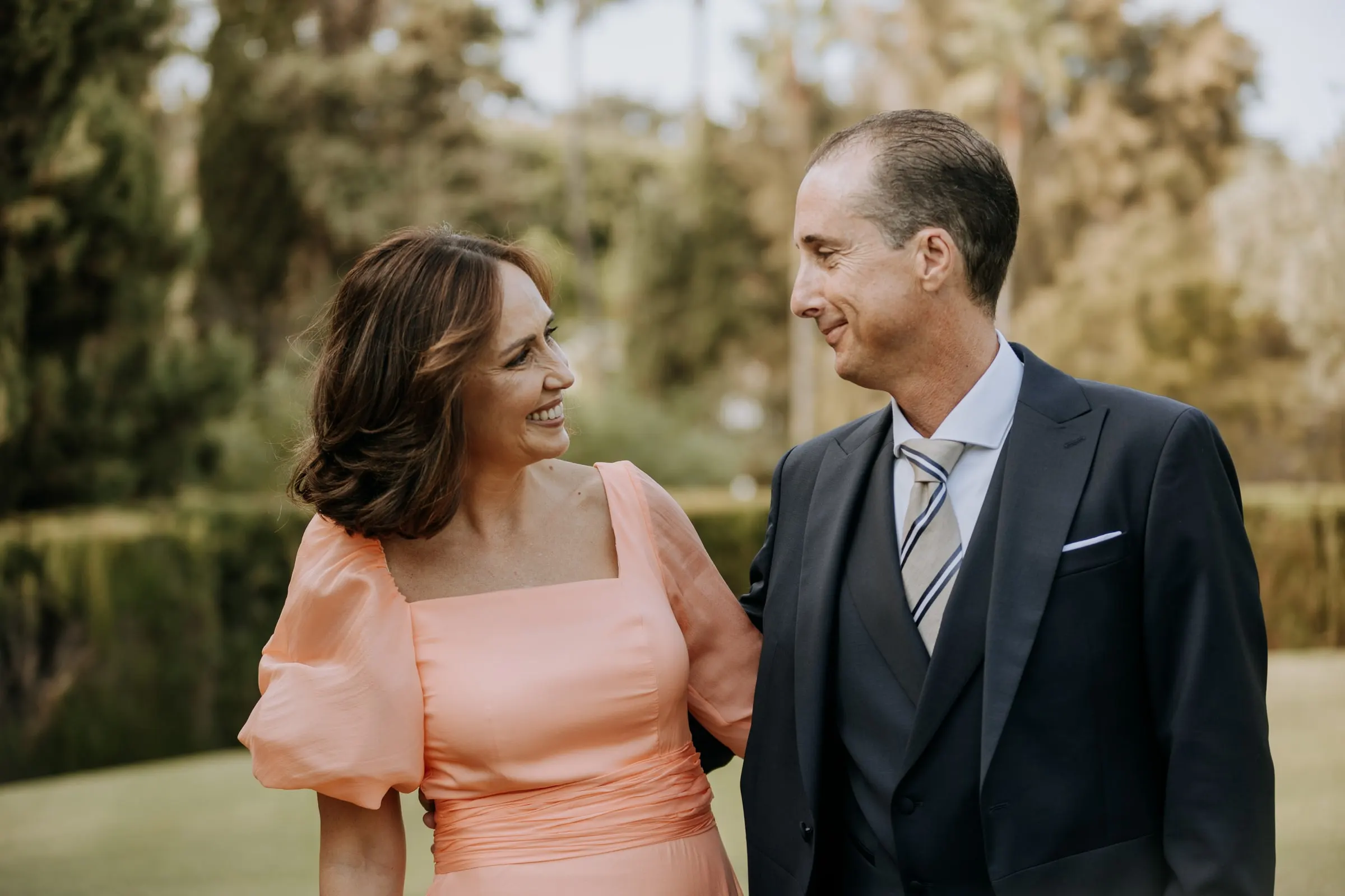 Woman in a peach dress and a man in a morning suit smiling at each other in the garden