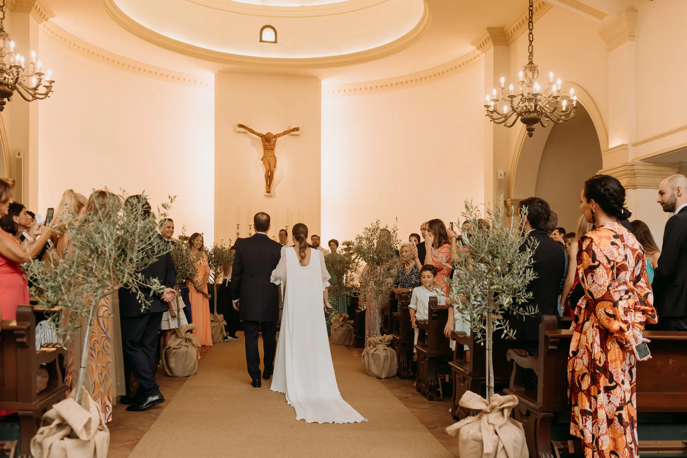 Bride walking up the aisle between potted olive trees towards the white apse and crucifix of Nuestra Señora de la Merced