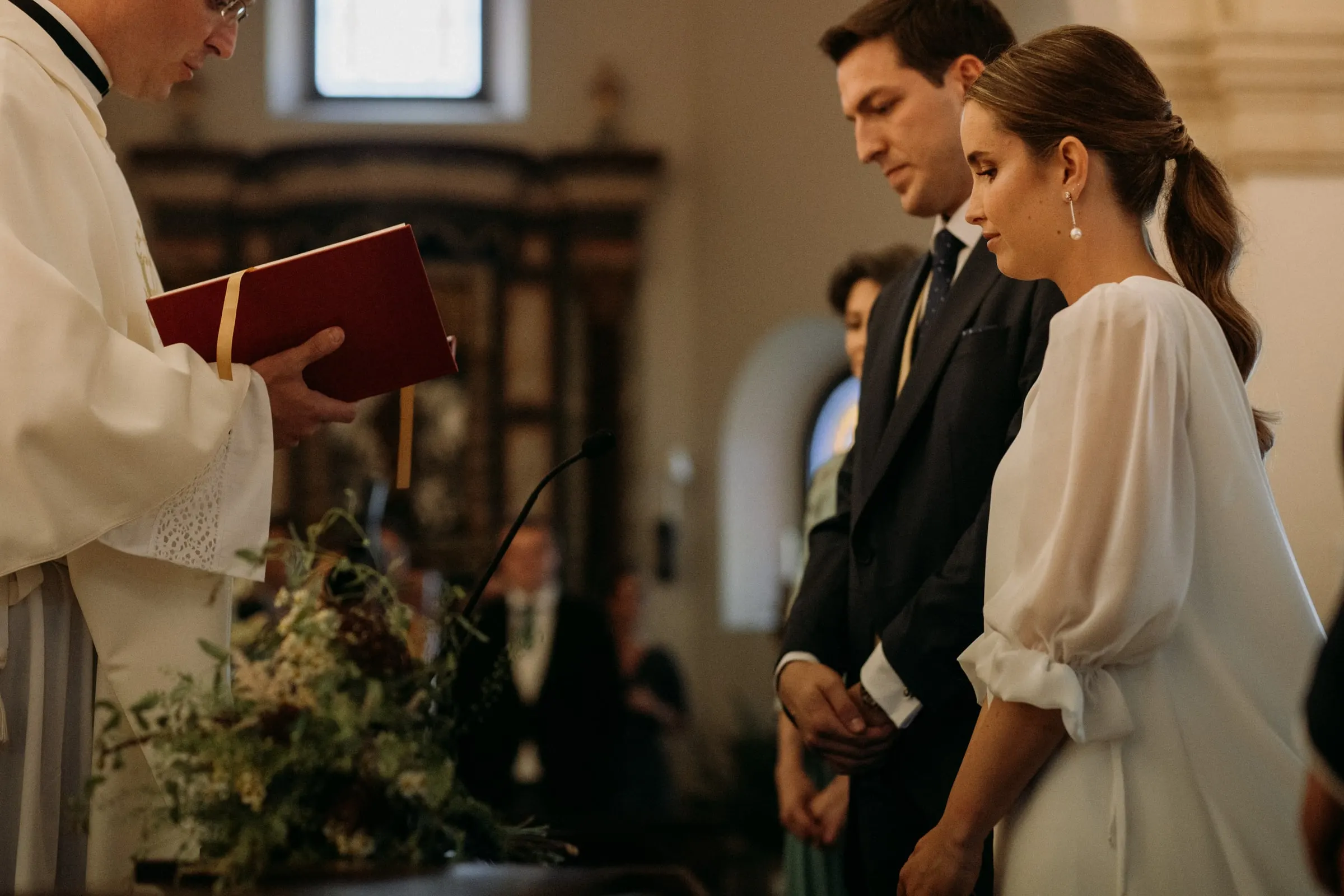 Priest reading from a red book as the bride and groom stand among olive branches at the altar