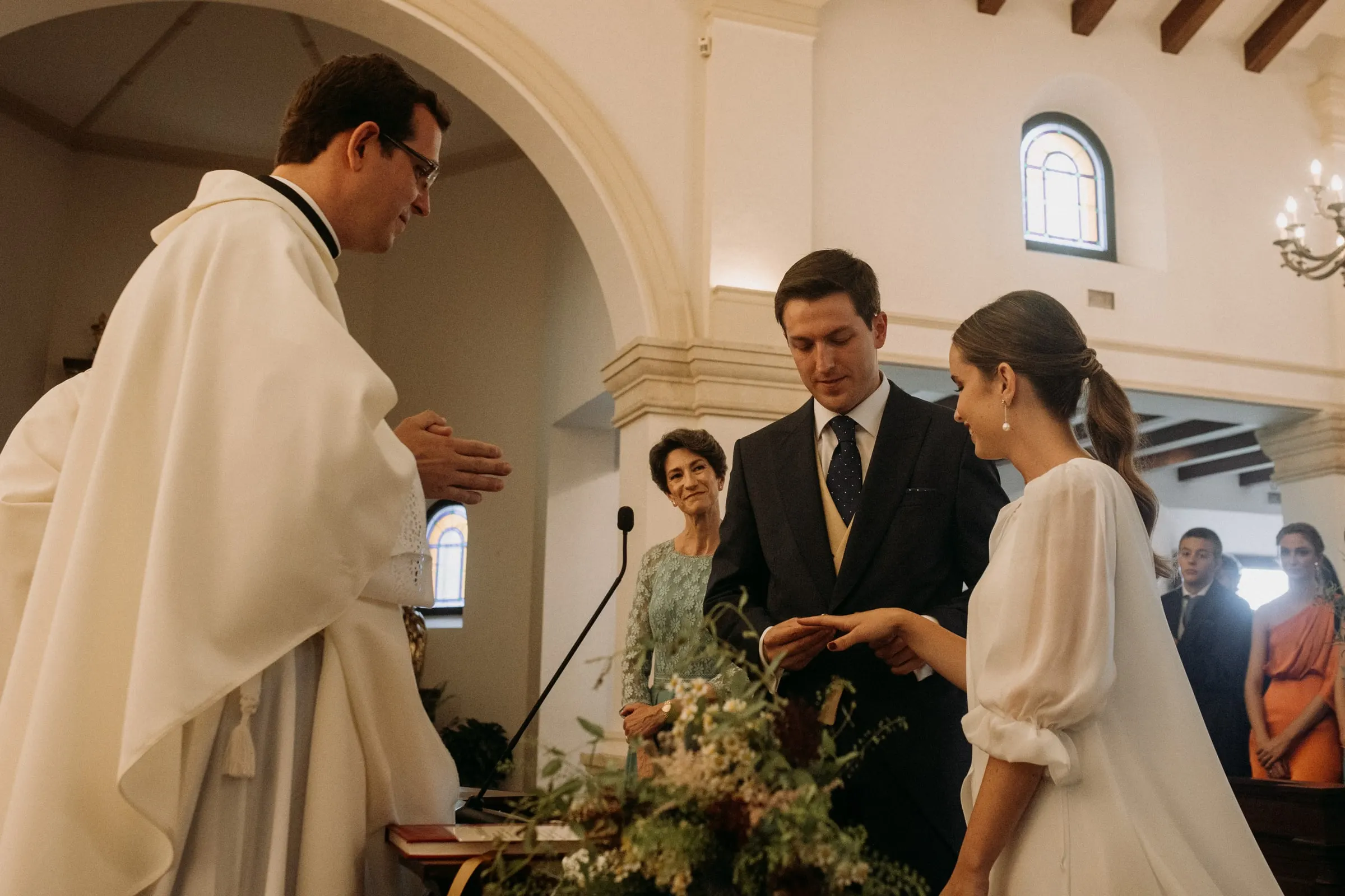 Priest in a cream chasuble speaking to the couple beside the altar flowers in the church in Sotogrande