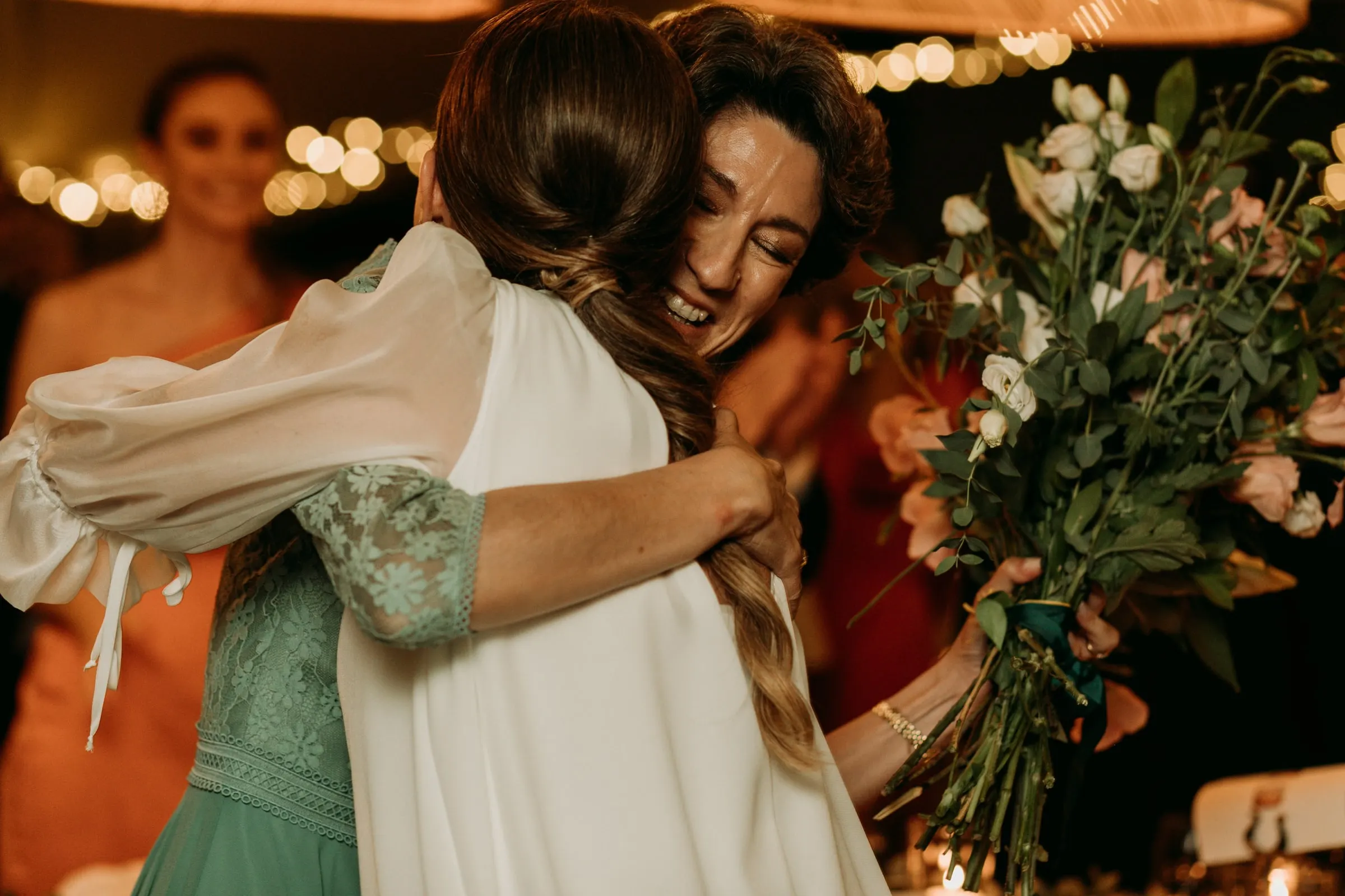 Bride hugging a guest in green, bouquet in hand, under the fairy lights