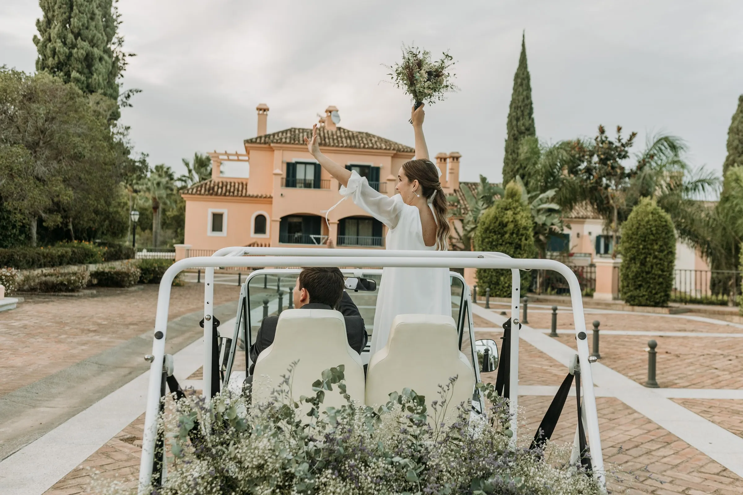 La novia de pie en el coche descapotable saluda con el ramo al alejarse por Sotogrande