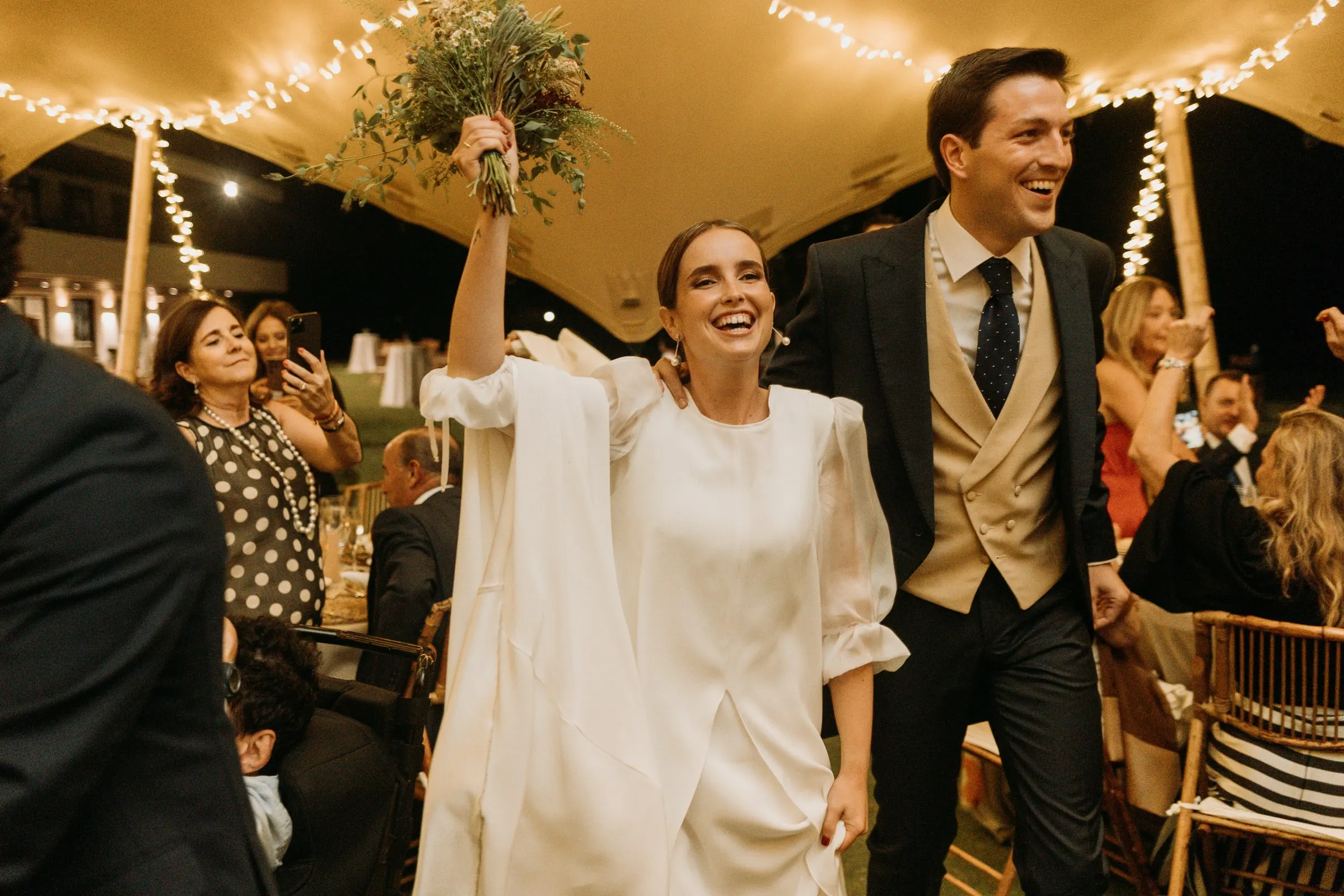 Bride raising her bouquet next to the groom under the fairy lights in Sotogrande
