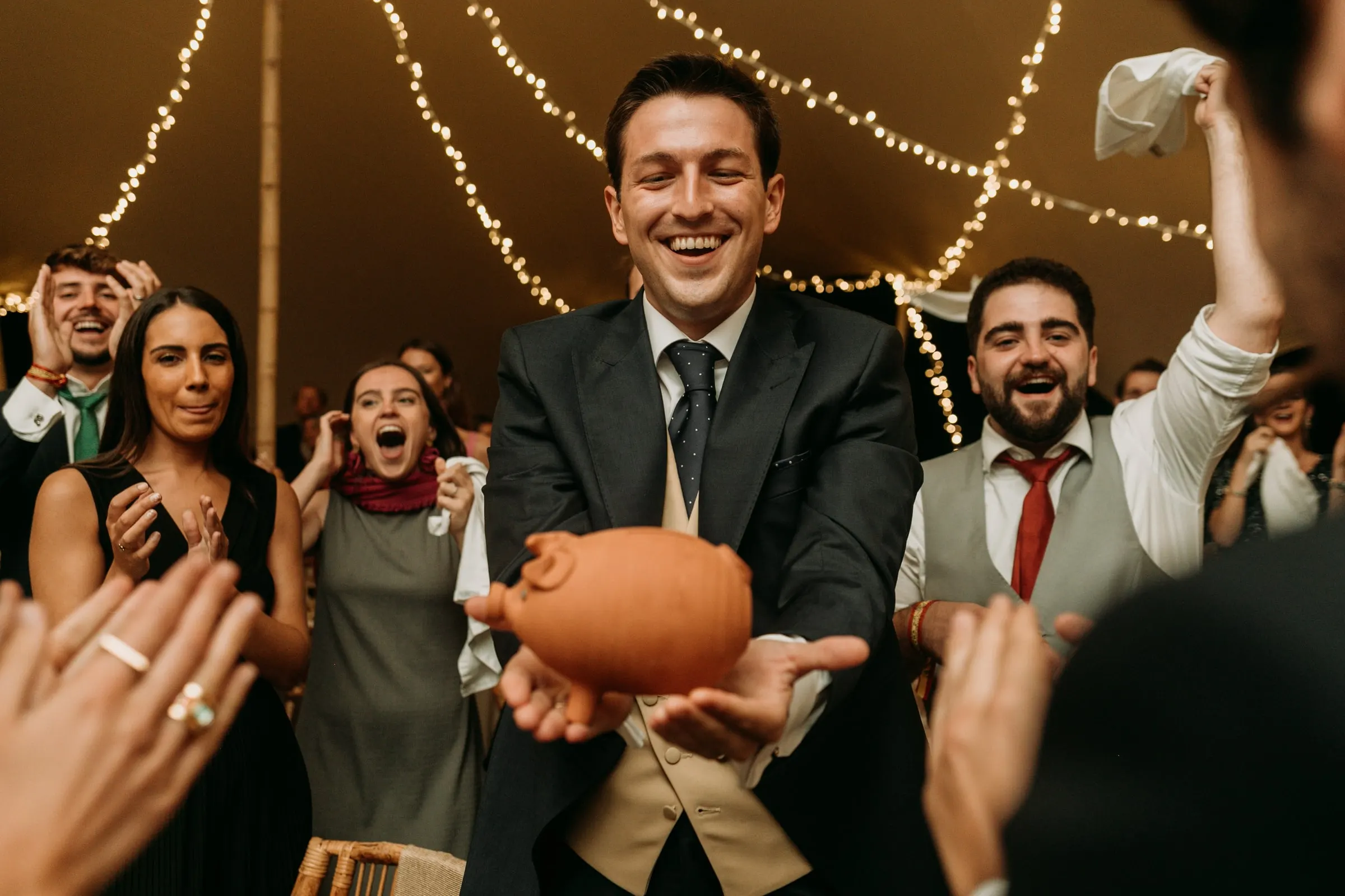 Groom holding a clay piggy bank as friends cheer around him
