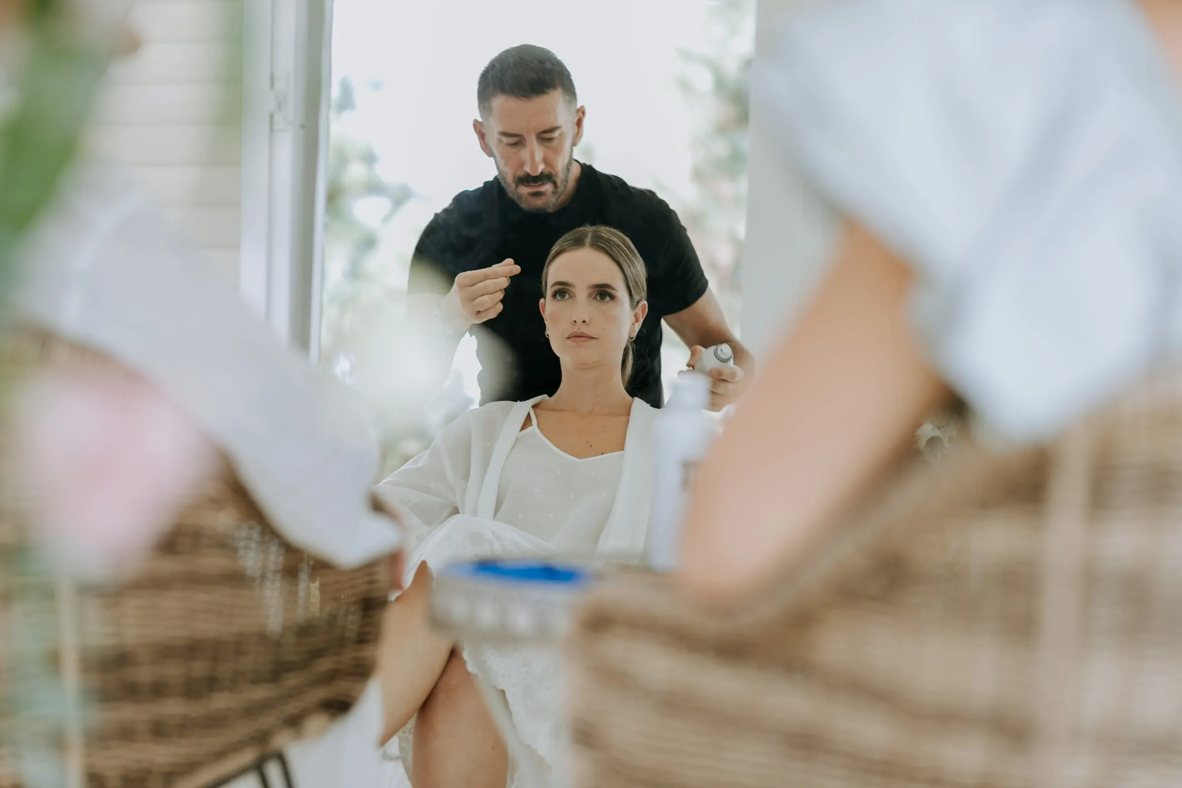 Bride in a wicker chair while a hairdresser in black works on her hair, seen between chairs and flowers