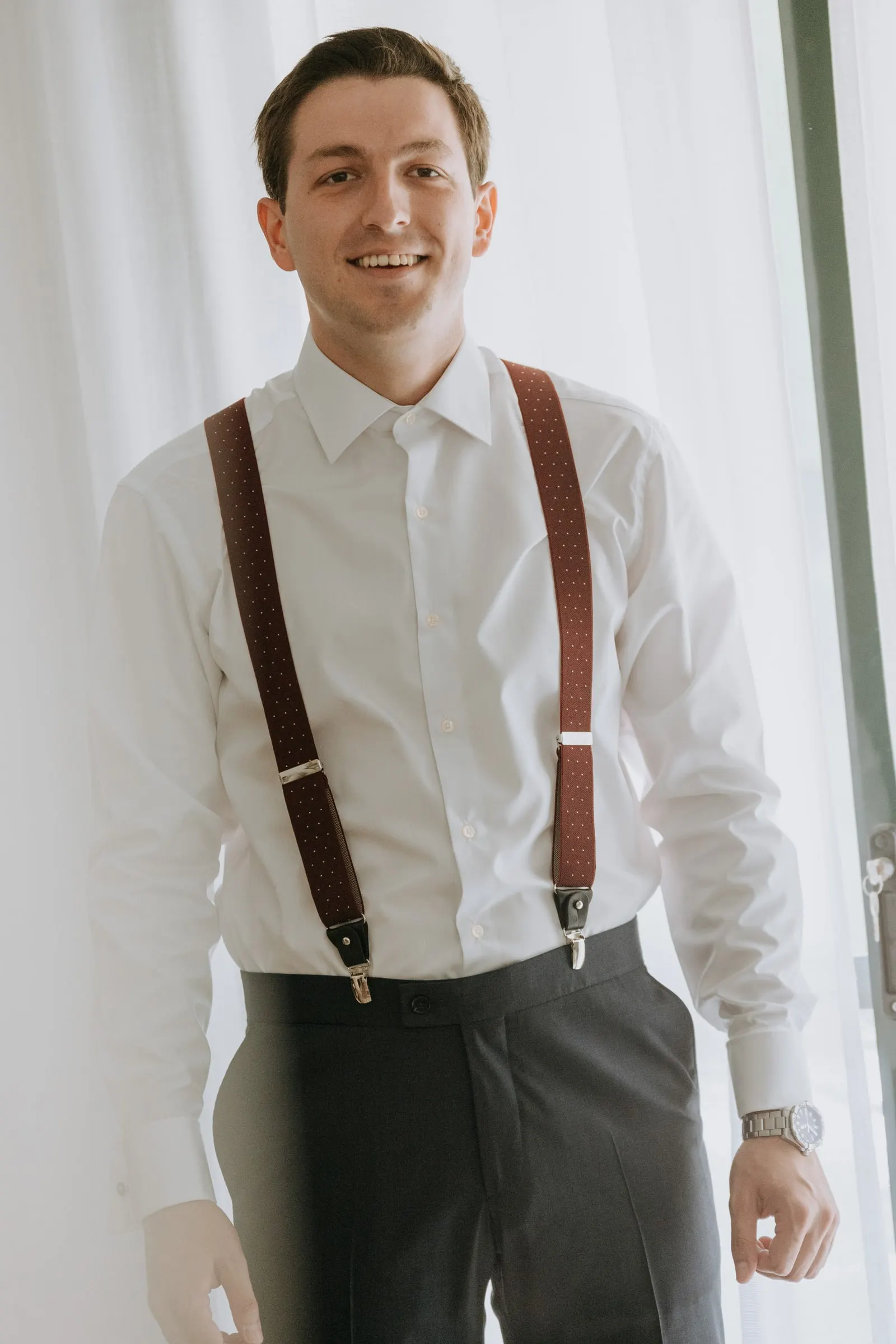 Groom smiling in his white shirt and burgundy braces by the window before the wedding in Sotogrande