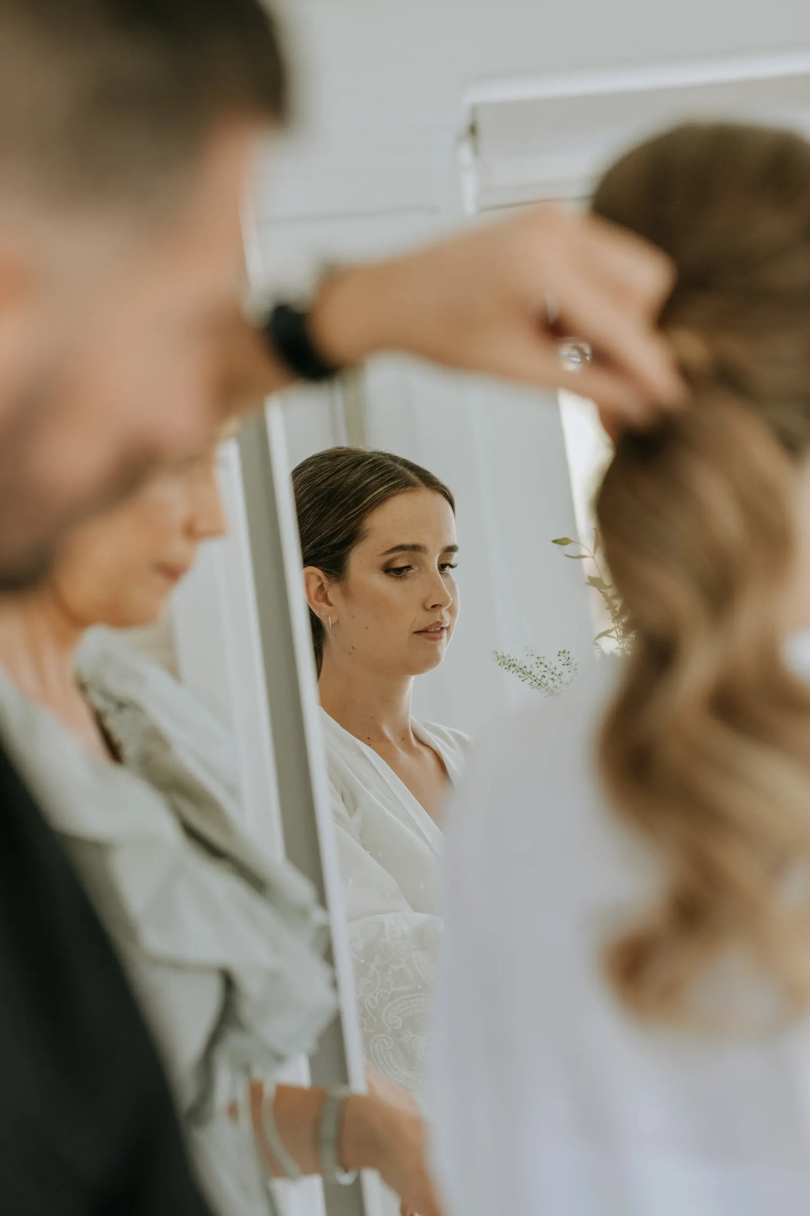 Bride seen in the mirror while the hairdresser finishes a guest's long curls