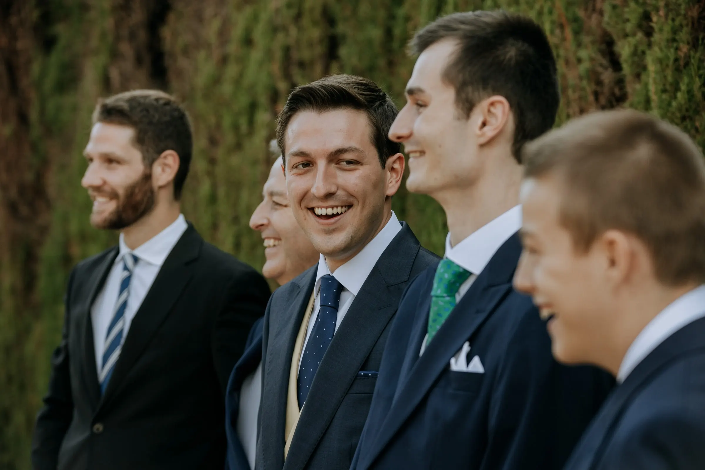 Close-up of the groomsmen laughing side by side in morning suits by the hedge