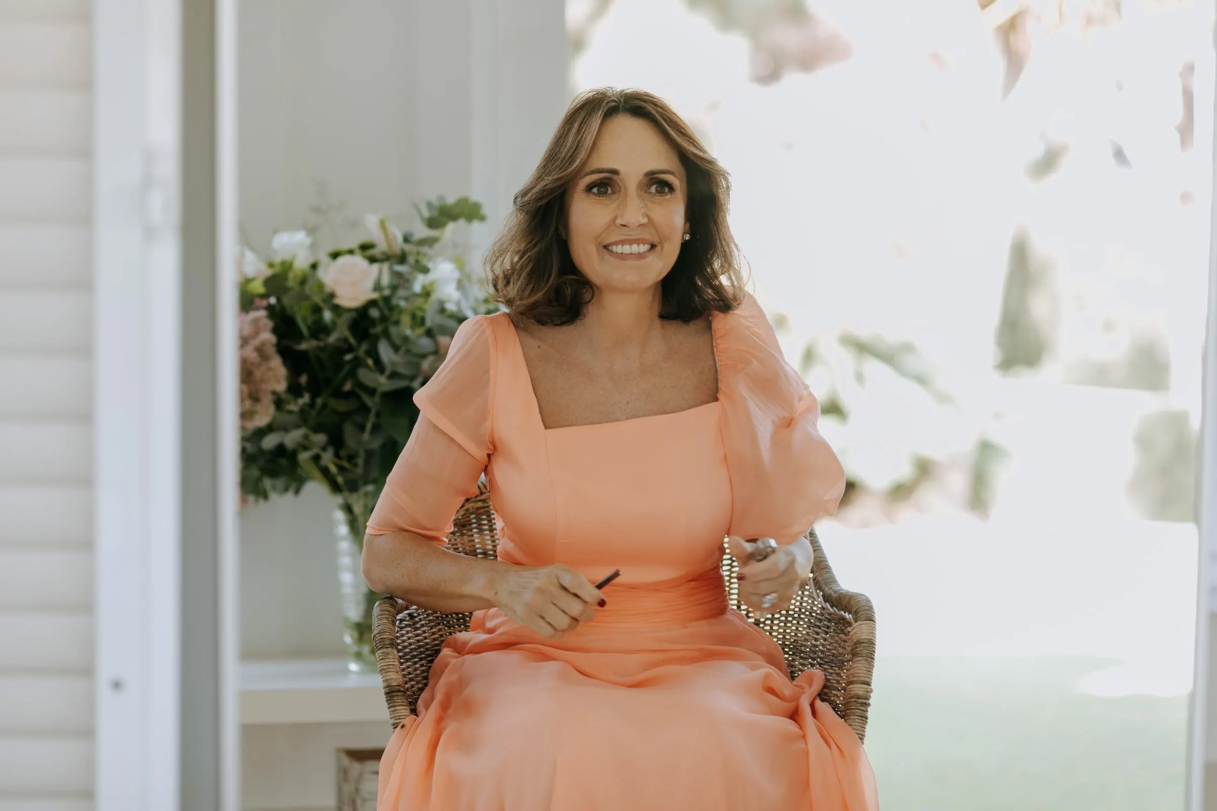 Woman in a peach dress smiling in a wicker chair by the open window