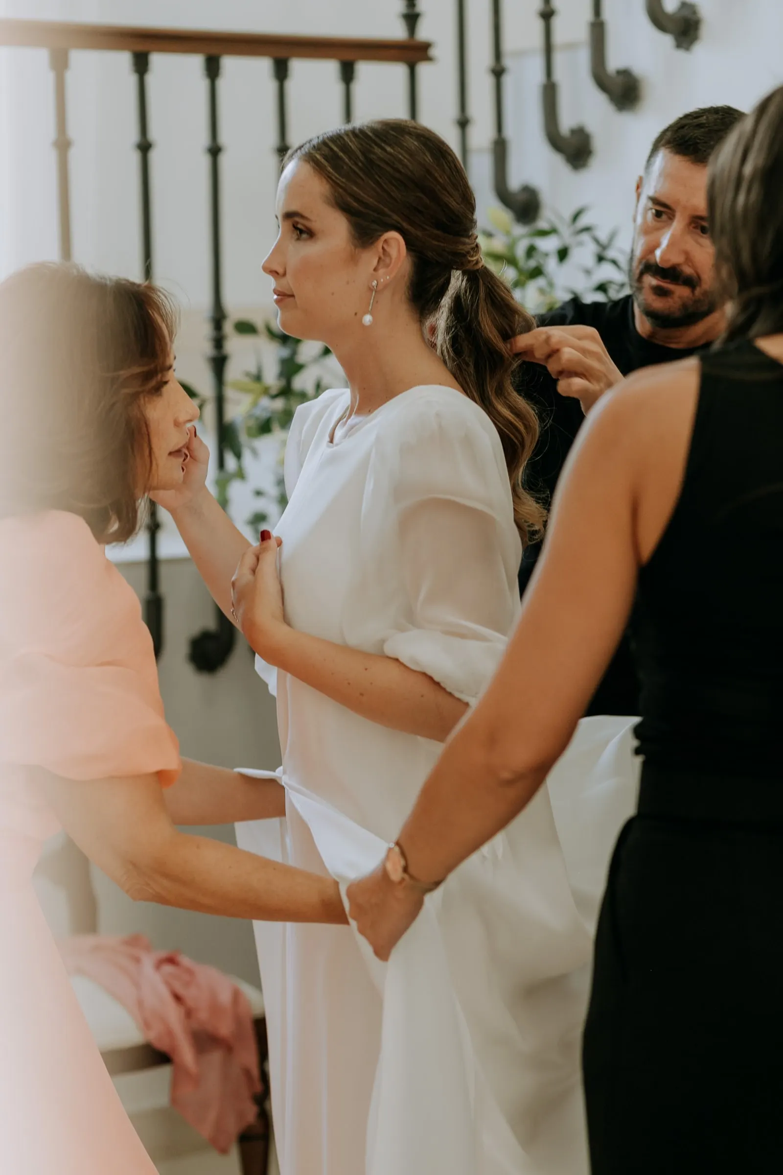 Bride surrounded by three women fixing her dress and make-up at the foot of the iron staircase