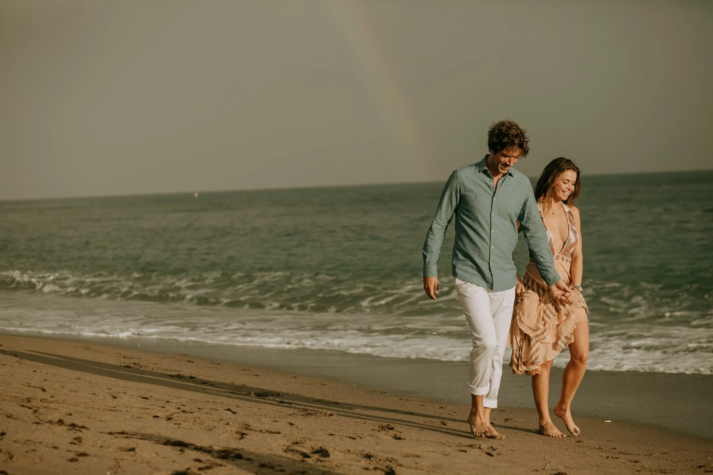 Lauren y Arnaud caminan descalzos por la orilla bajo un arcoíris tenue, sesión preboda en la playa de Sotogrande