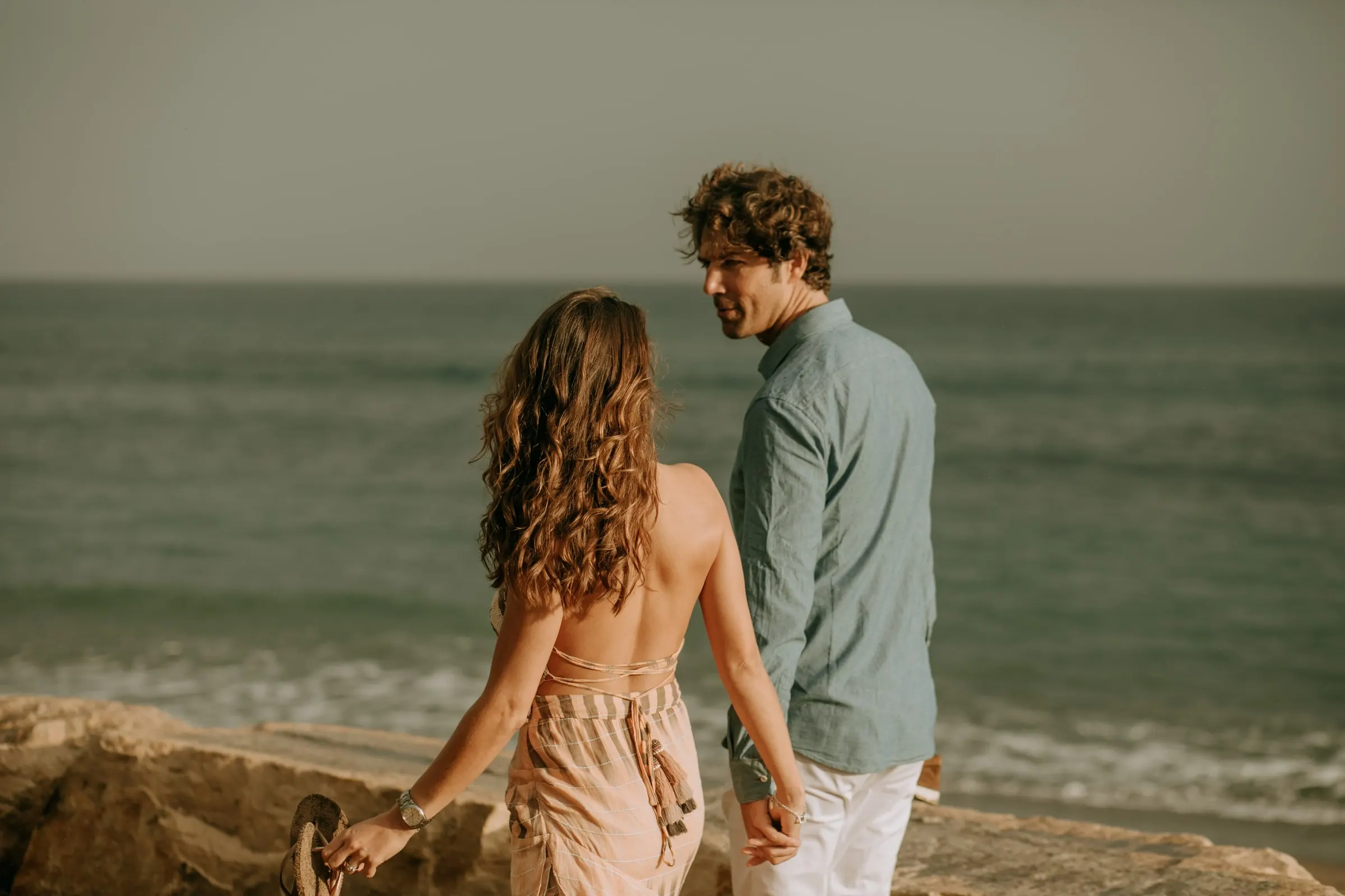 La futura novia, con vestido rosa de espalda abierta, va hacia su prometido por las rocas, playa de Sotogrande