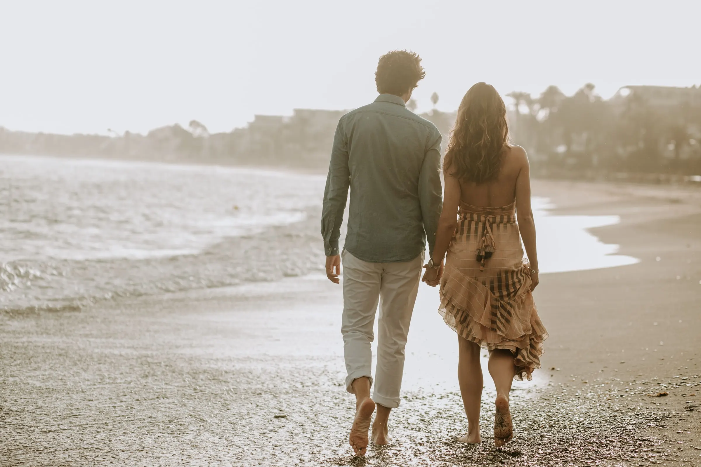 La pareja, de espaldas, camina descalza por la orilla hacia la luz, sesión preboda en la playa de Sotogrande