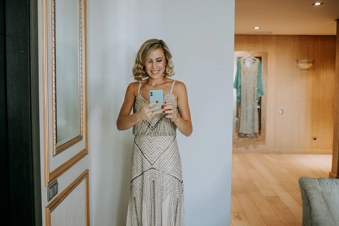 Una invitada con vestido de abalorios se hace un selfi en el espejo, con el vestido de la novia al fondo