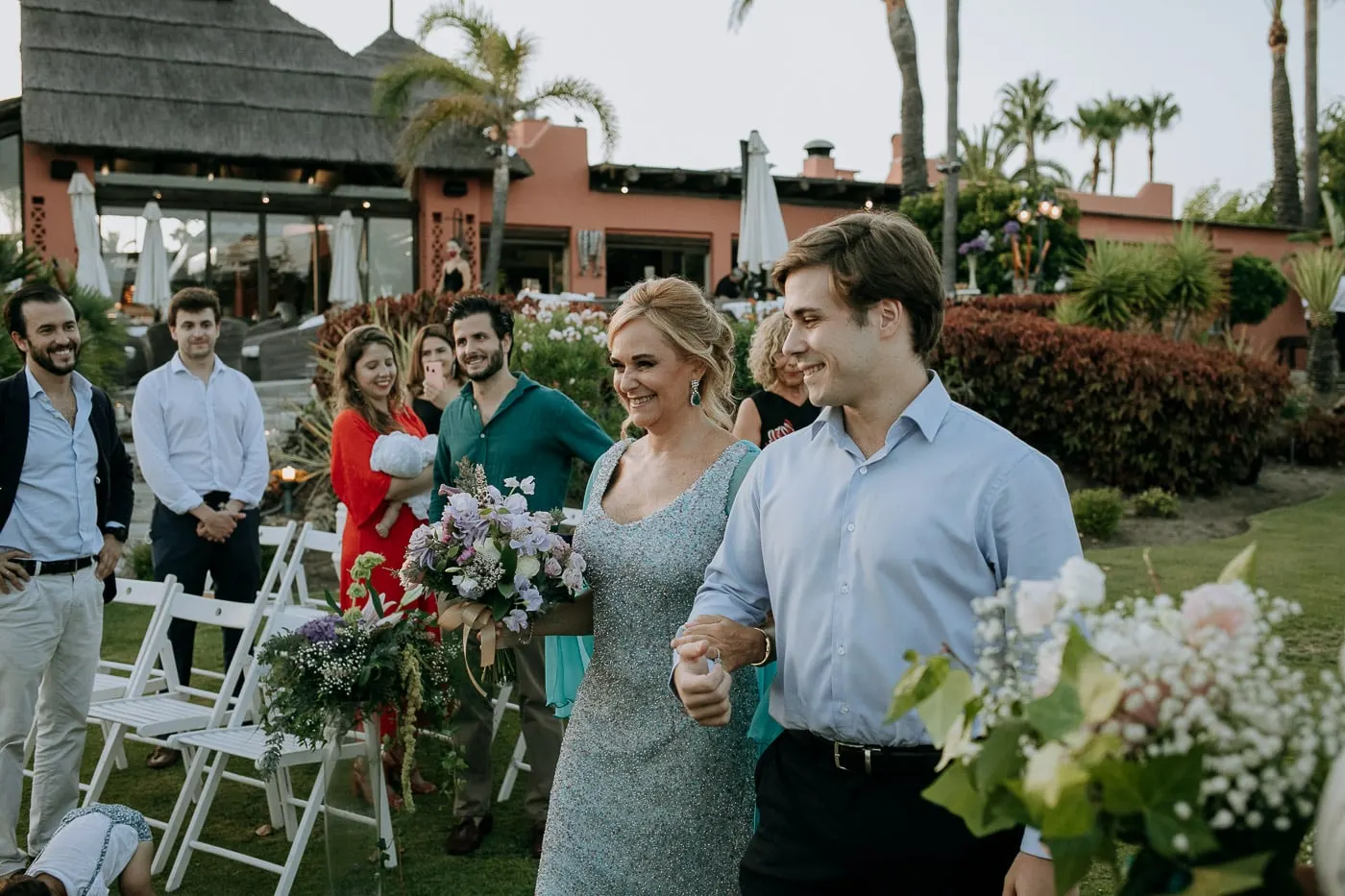 La novia entra en la ceremonia con el restaurante de tejado de paja detrás, Tikitano