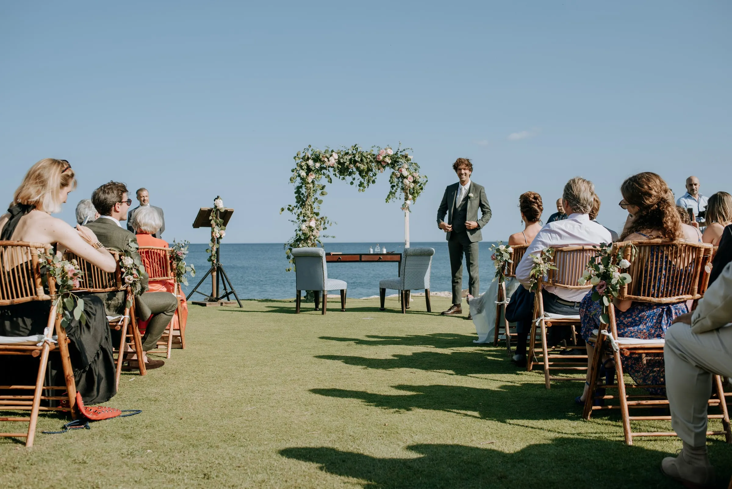 Invitados sentados en el césped mientras el novio espera junto al arco frente al mar, Trocadero, Sotogrande