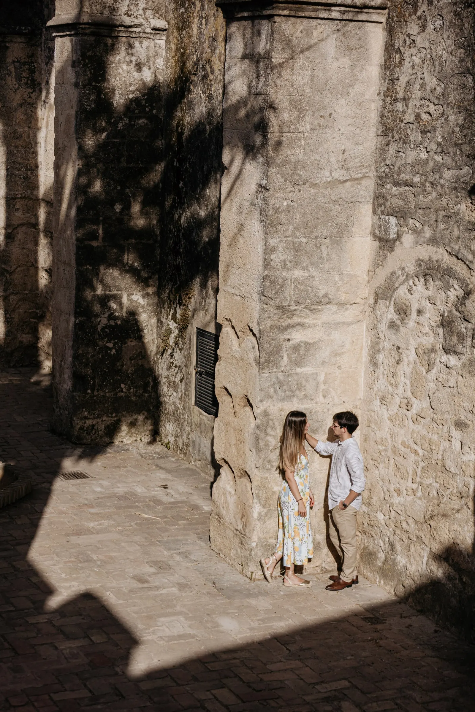 La pareja, pequeña ante altos muros de piedra entre luz y sombra profunda en Vejer