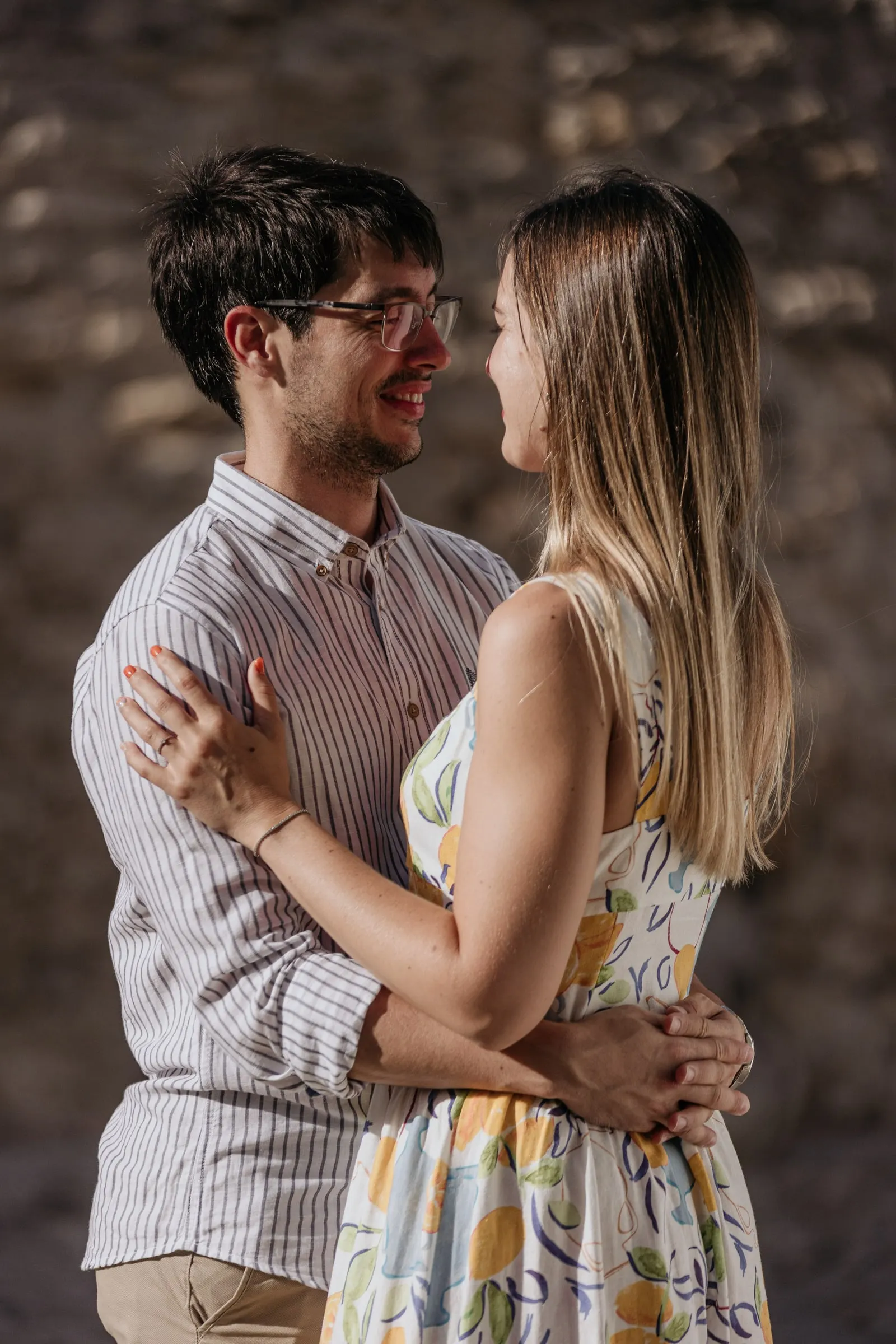 La futura novia con vestido de flores sonríe a su prometido junto a un muro de piedra en Vejer de la Frontera