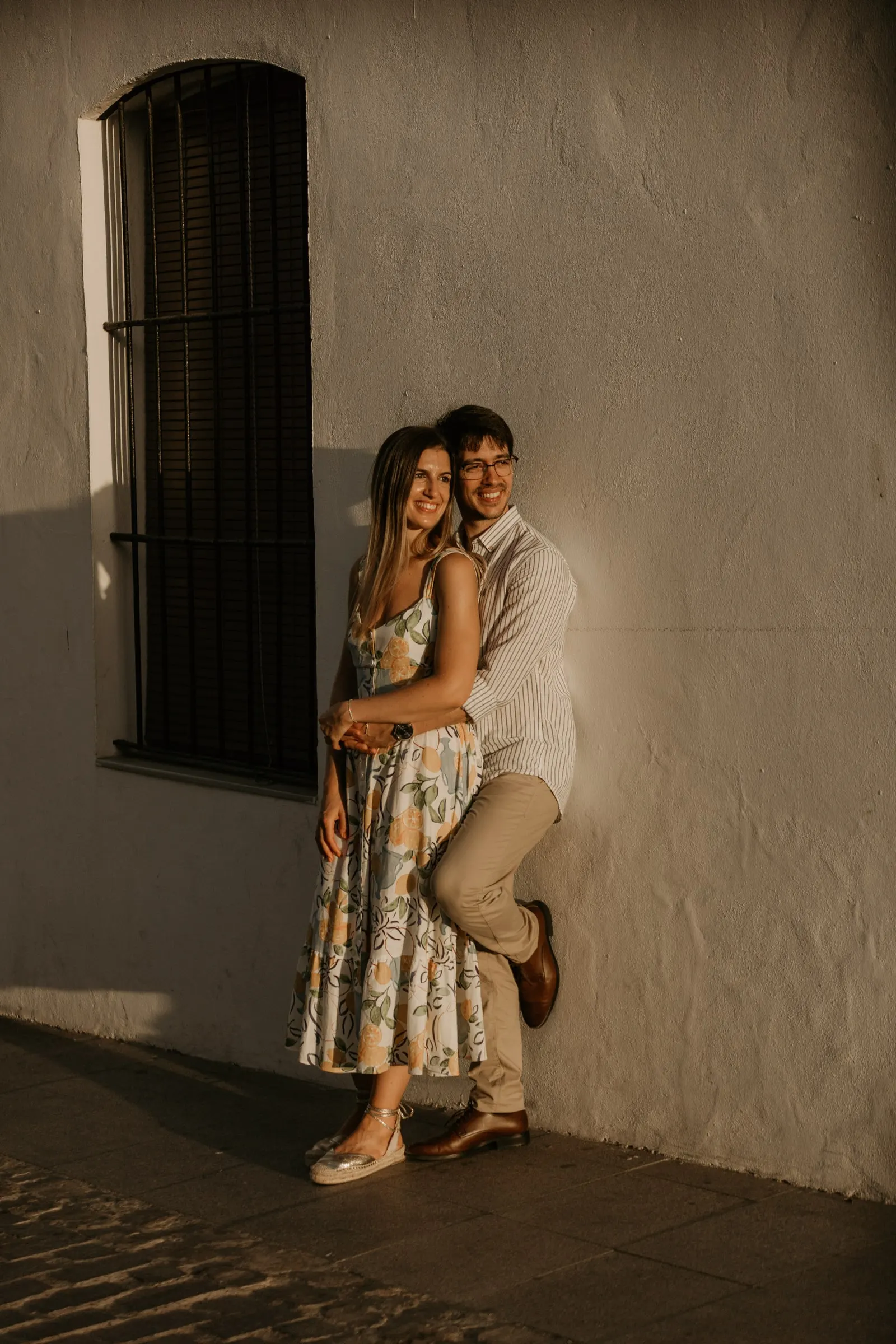 La pareja abrazada contra una pared blanca junto a una ventana oscura con la luz de la tarde, Vejer