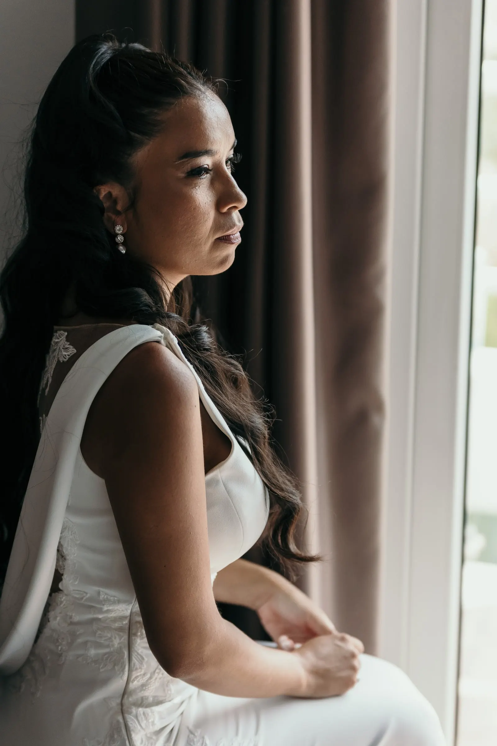 La novia, ya vestida, sentada junto a la ventana mira hacia fuera en silencio