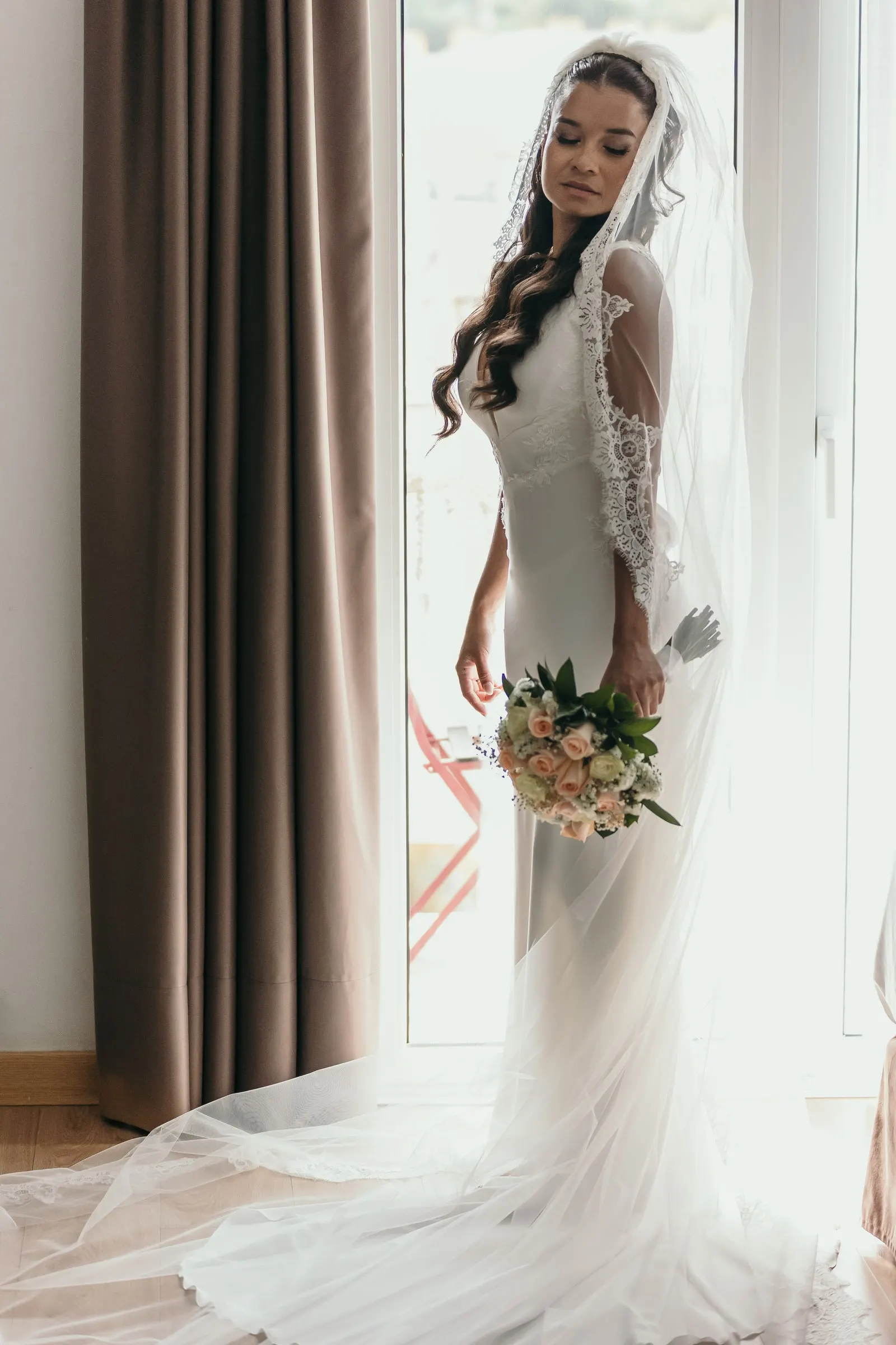 La novia de pie con velo y ramo de rosas rosas junto a un ventanal, Marbella