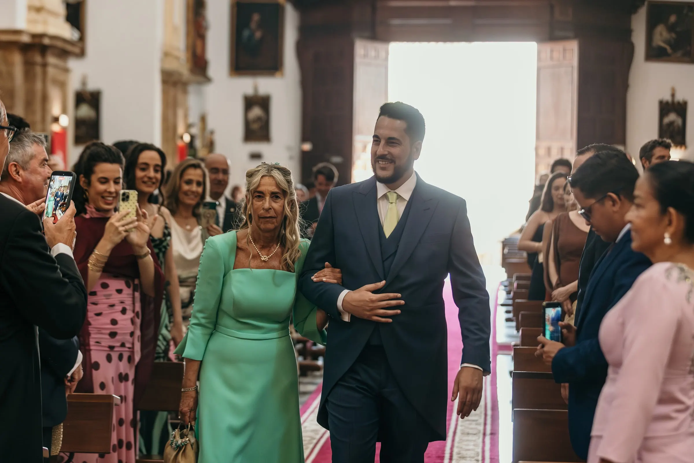 El novio entra por el pasillo de la iglesia del brazo de su madre, de vestido verde agua