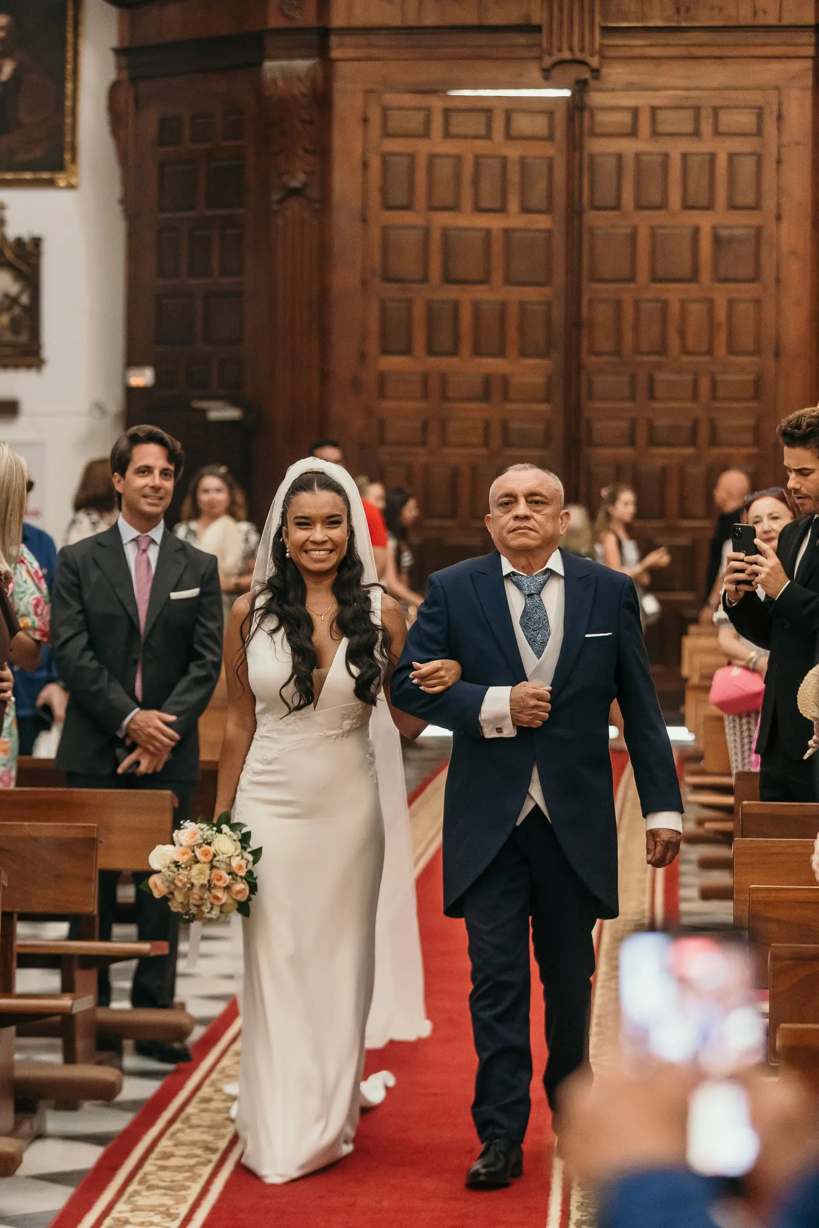 La novia avanza por la alfombra roja del brazo de su padre mientras los invitados hacen fotos, boda en iglesia de Marbella