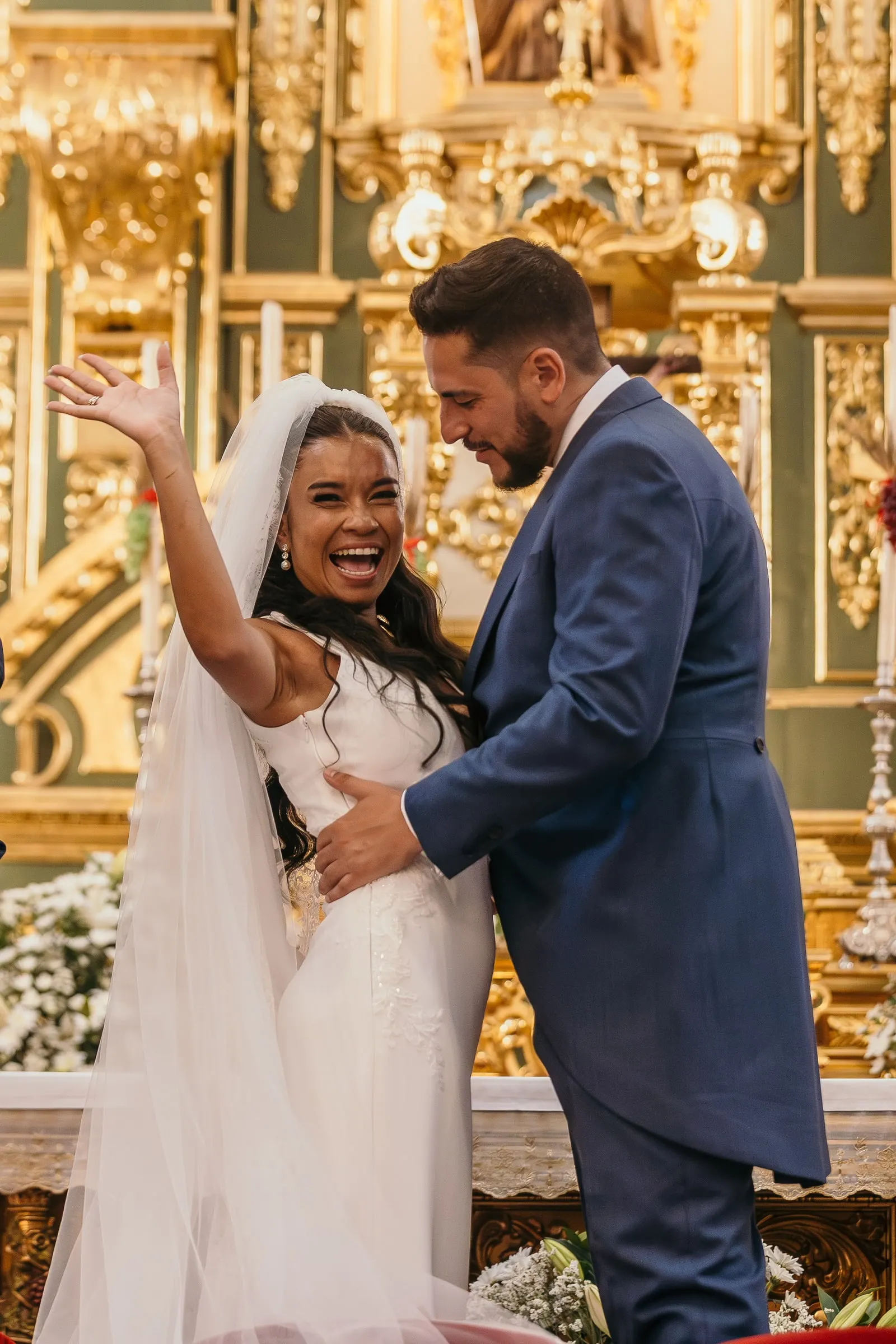 La novia levanta el brazo entre risas mientras el novio la abraza en el altar