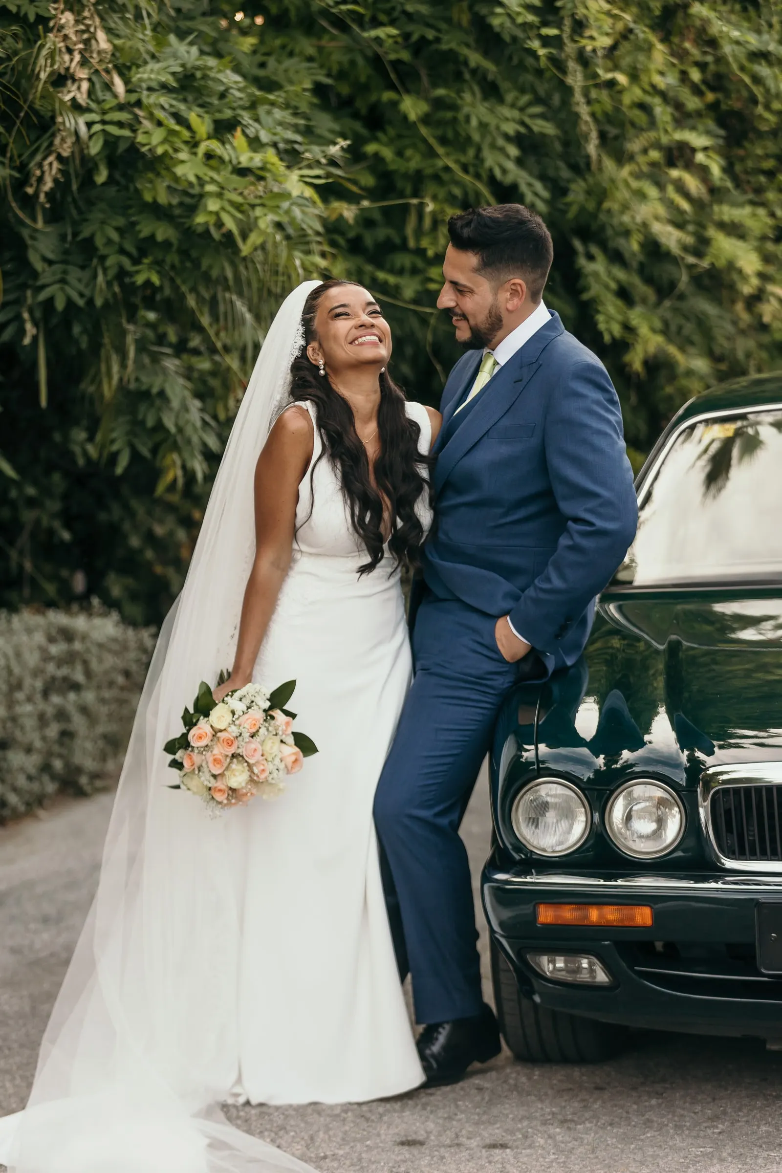 Couple laughing and leaning against the vintage Jaguar, bride holding her rose bouquet