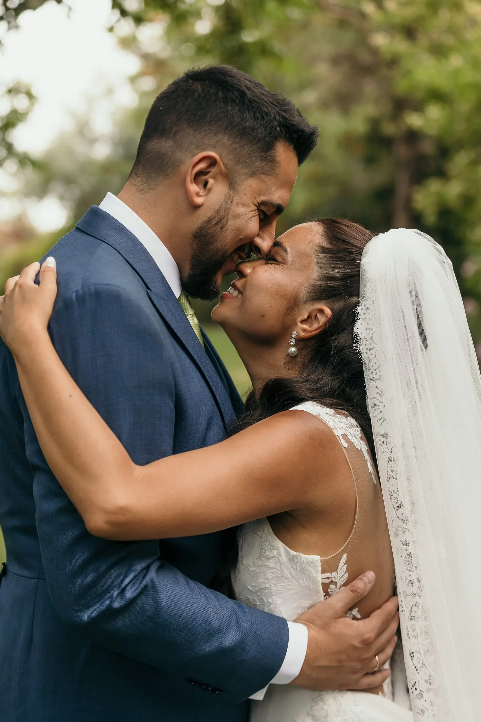 Groom kissing the bride's cheek in a tight embrace under the trees