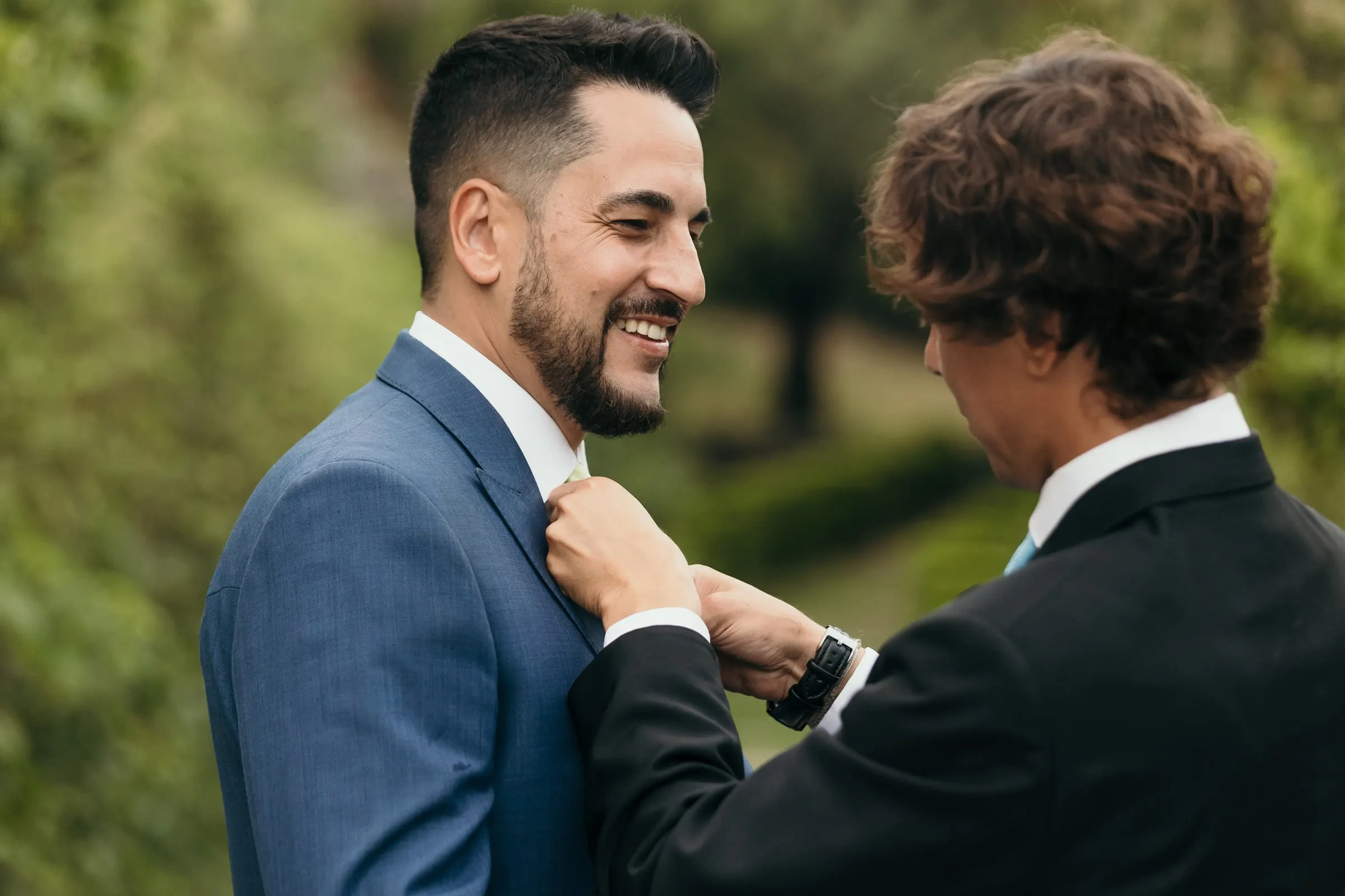 Un invitado joven le recoloca la corbata al novio en el jardín, que sonríe